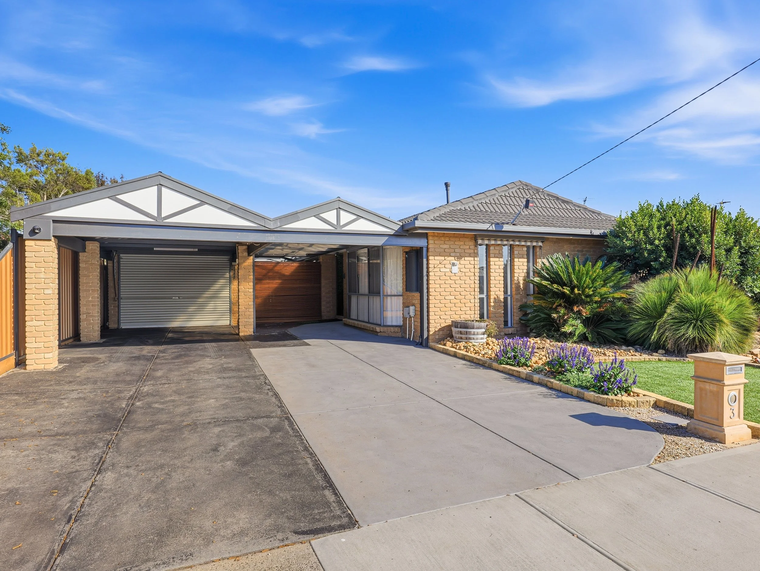 A suburban house with a garage, brick walls, a tiled roof, and a small front garden with purple flowers, a mailbox, and green bushes, under a clear blue sky.