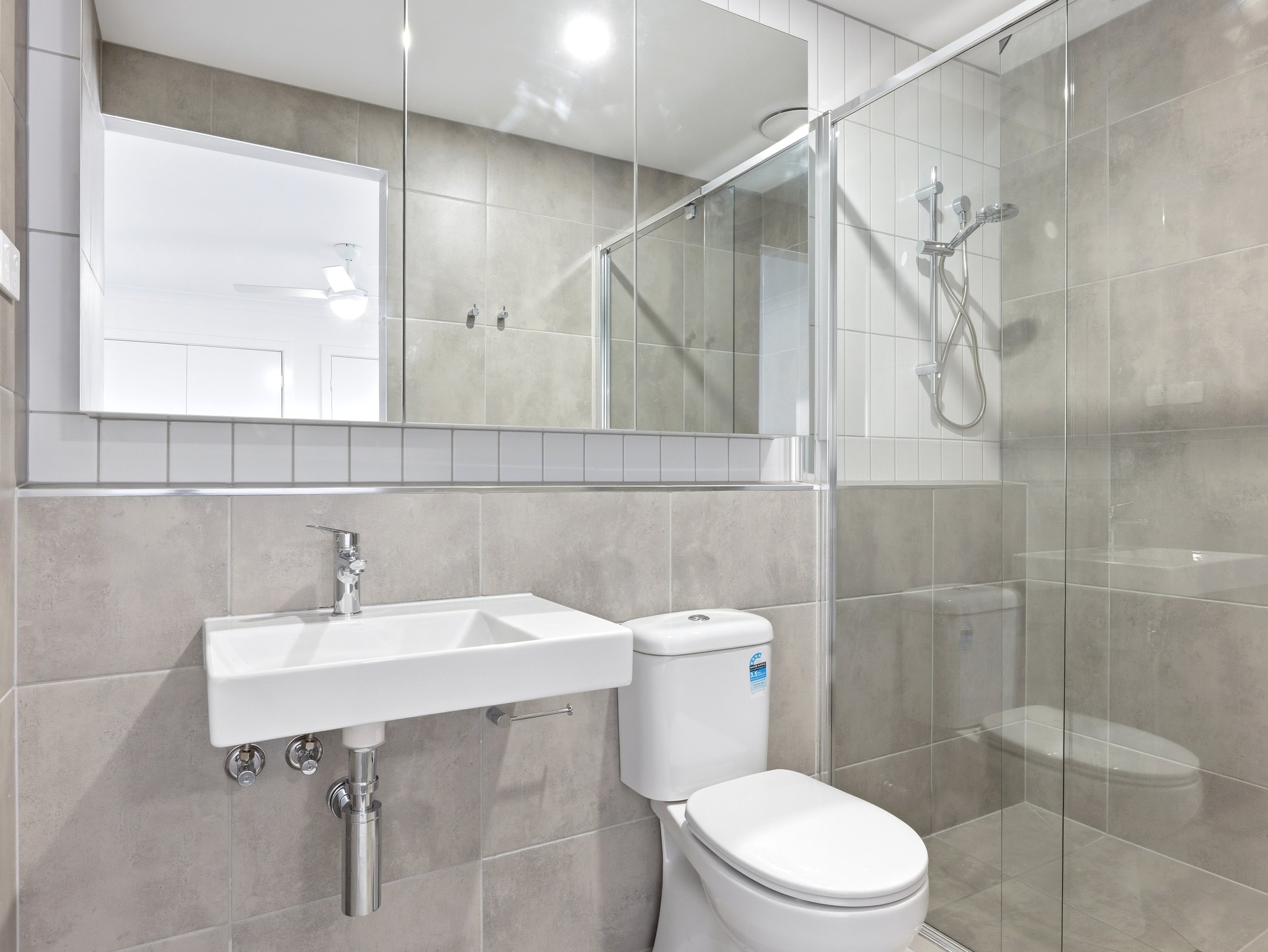 A small bathroom with a mirror, sink, toilet, and shower with glass doors, tiled walls, and a ceiling light.