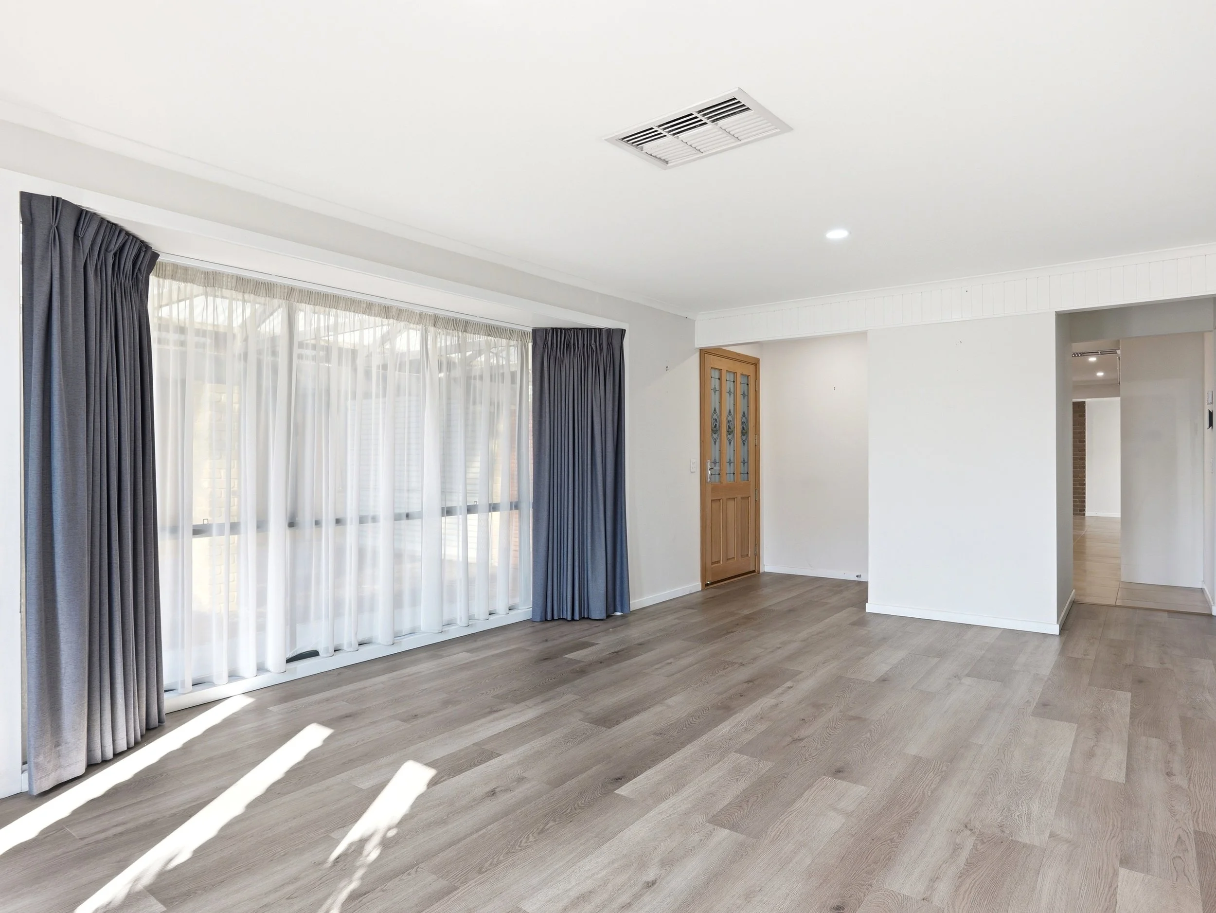 Bright living room with large windows, dark curtains, a wooden door, and a white ceiling with a vent