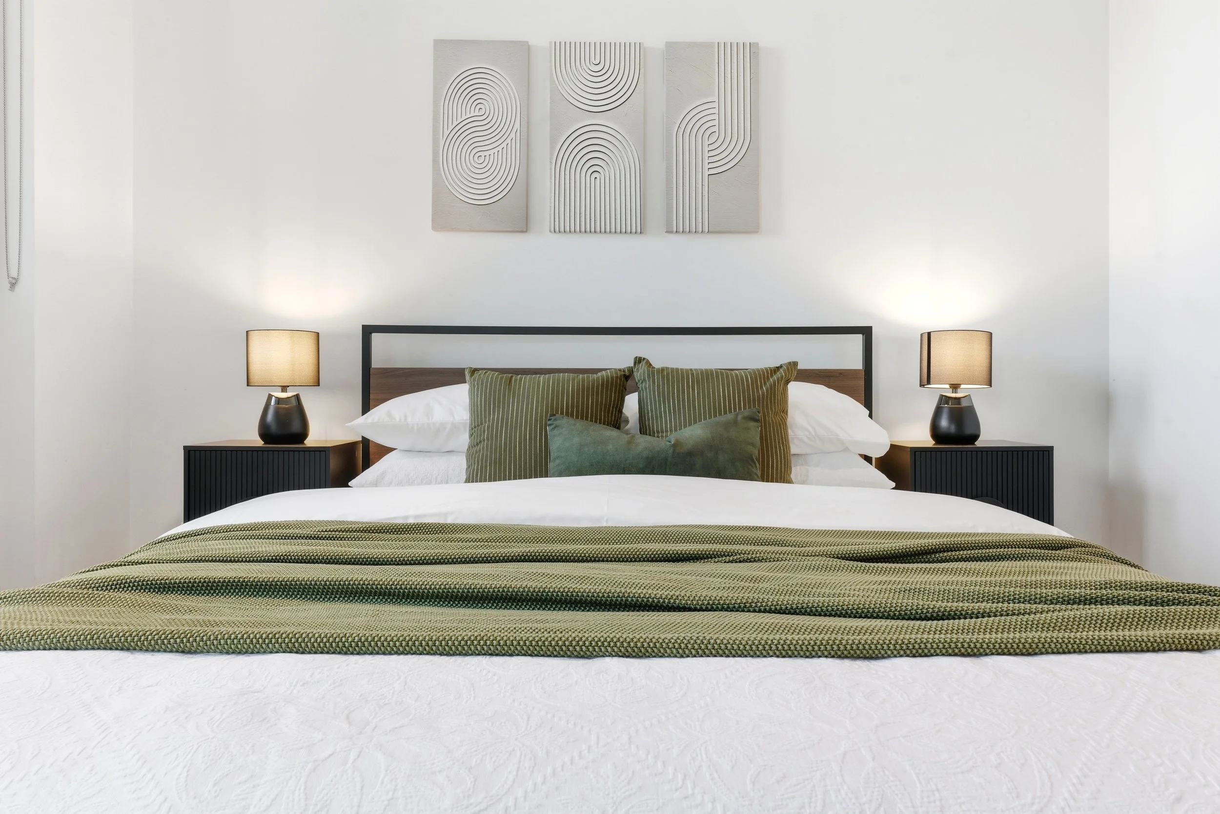 A neatly made bed with a white quilt, green and striped pillows, and a textured green bedspread, flanked by black nightstands with beige lamps, and decorated with modern gray wall art above.