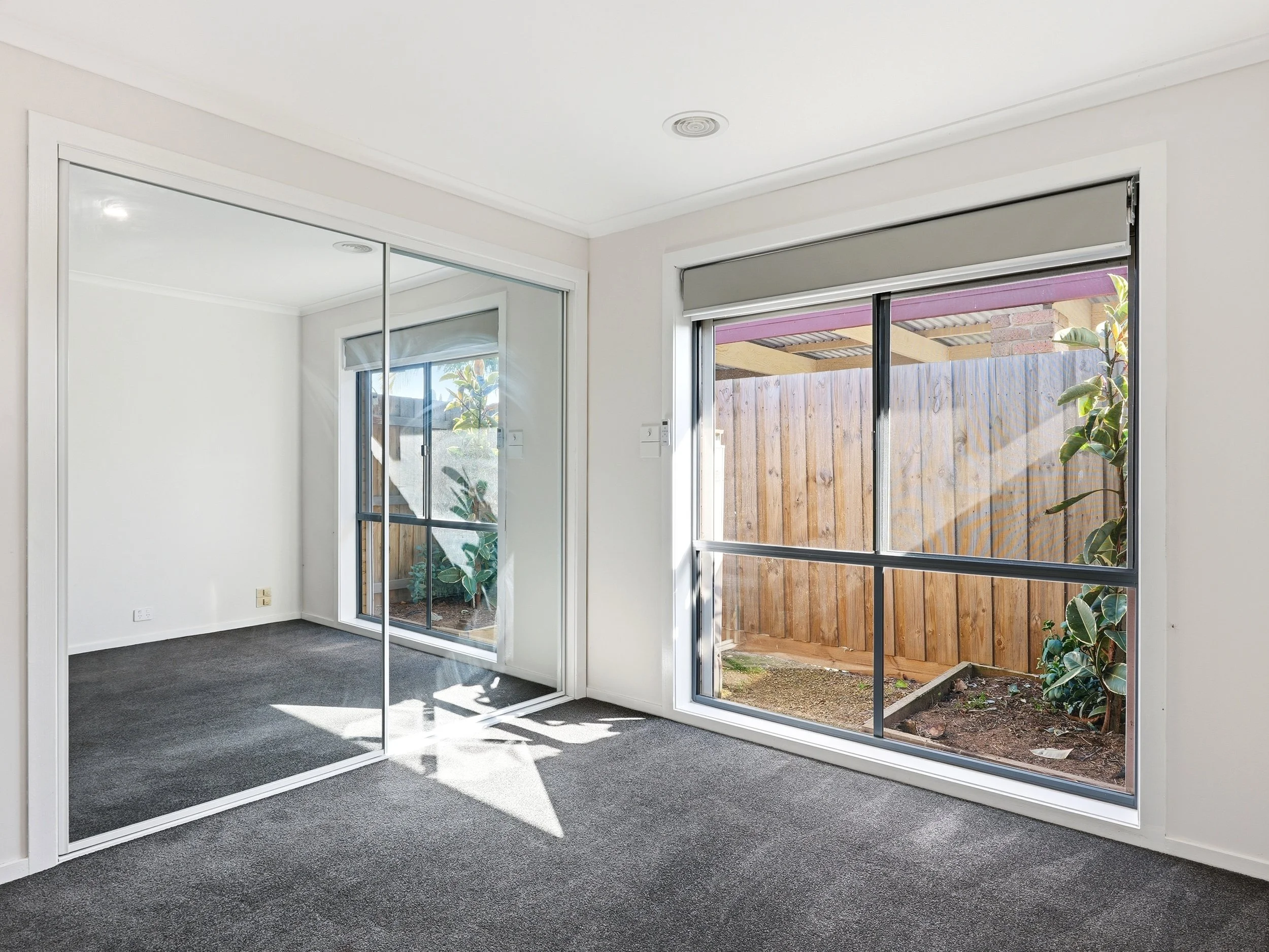 A room with large windows, a sliding mirrored closet door, and a view of a backyard with a wooden fence and plants.