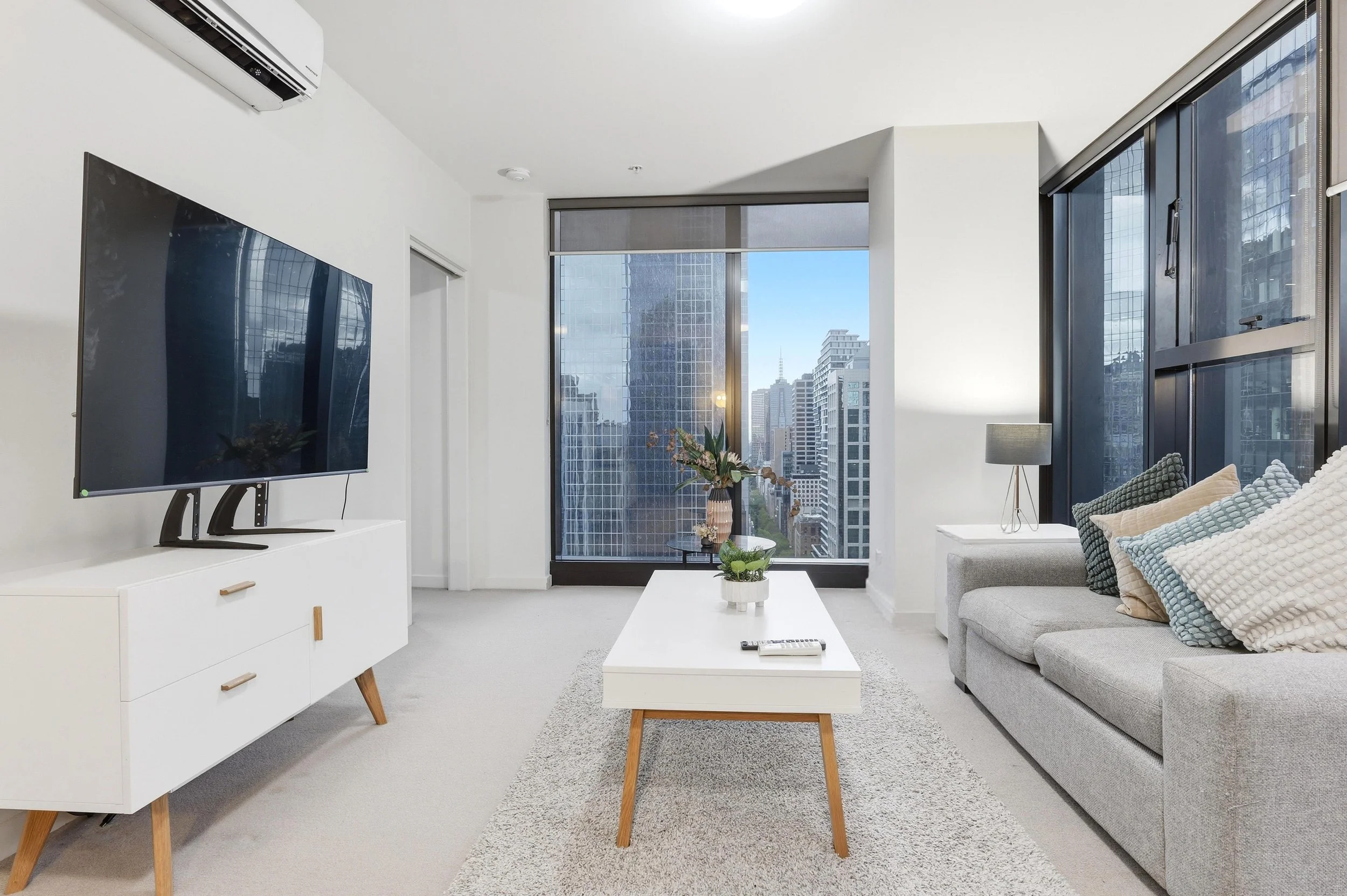 Modern living room with large windows, a gray sofa with pillows, a white coffee table with a remote control, and a white media console with a TV. Cityscape view with tall buildings outside.