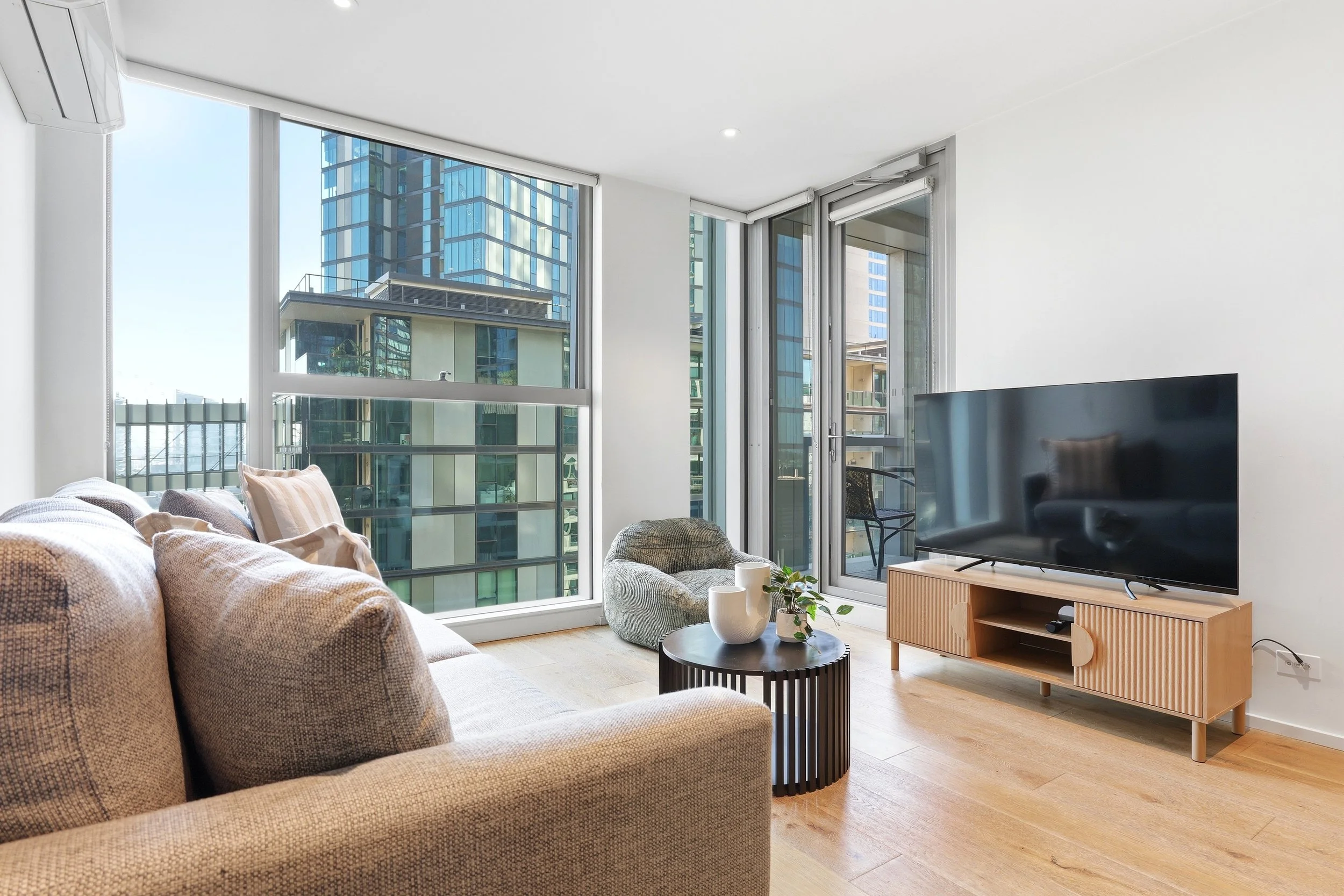 A modern living room with large windows showcasing city buildings. The room has a beige sofa with pillows, a gray armchair, a black coffee table with two white vases and plants, and a TV on a wooden stand. Light wood flooring and white walls complete