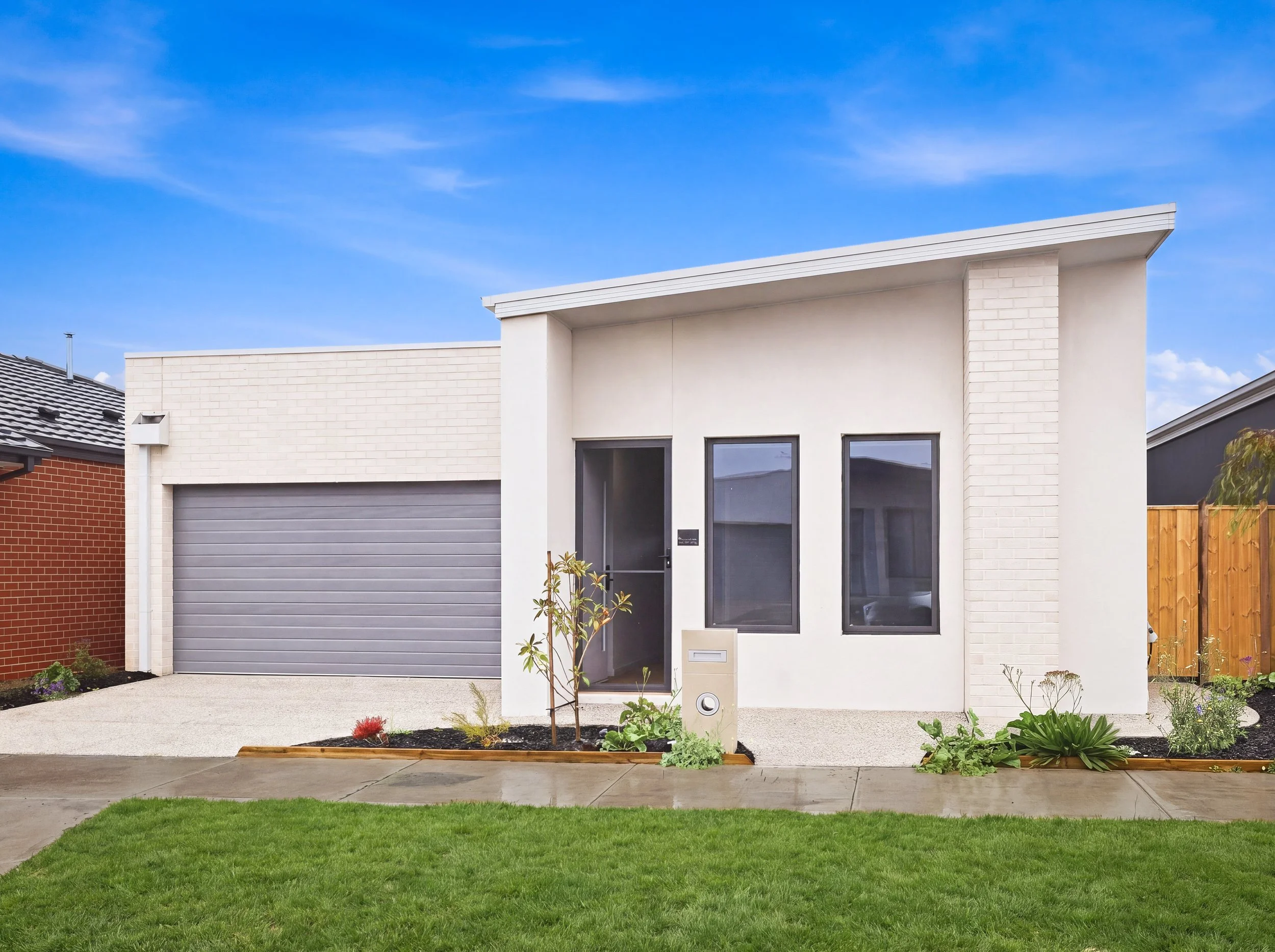Modern white house with large windows, a gray garage door, a small garden bed with plants, and a well-maintained lawn under a blue sky.