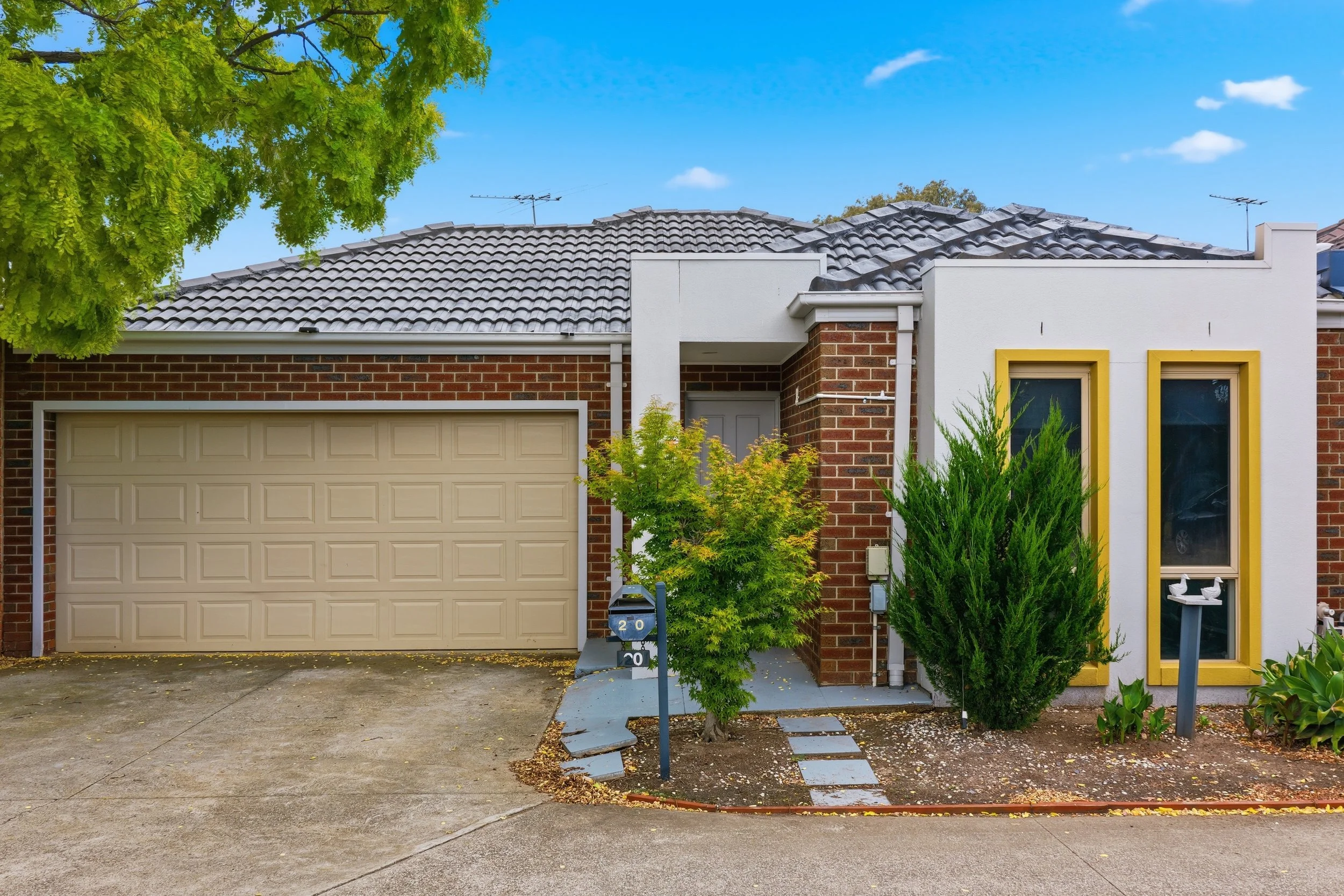 Front view of a modern suburban house with a beige garage door, white walls accented with yellow frames, and a brown brick facade. There are small trees and shrubs along the pathway leading to the front door, and a mailbox with the number 20. The driveway and sidewalk are visible, with some fallen leaves.
