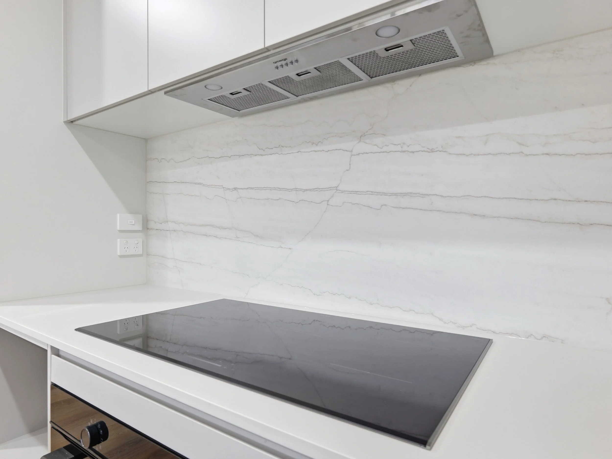A modern kitchen with a white marble countertop and a black electric stovetop. The backsplash features white marble with gray veining, and white upper cabinets are visible. There are electrical outlets and a control switch on the wall.