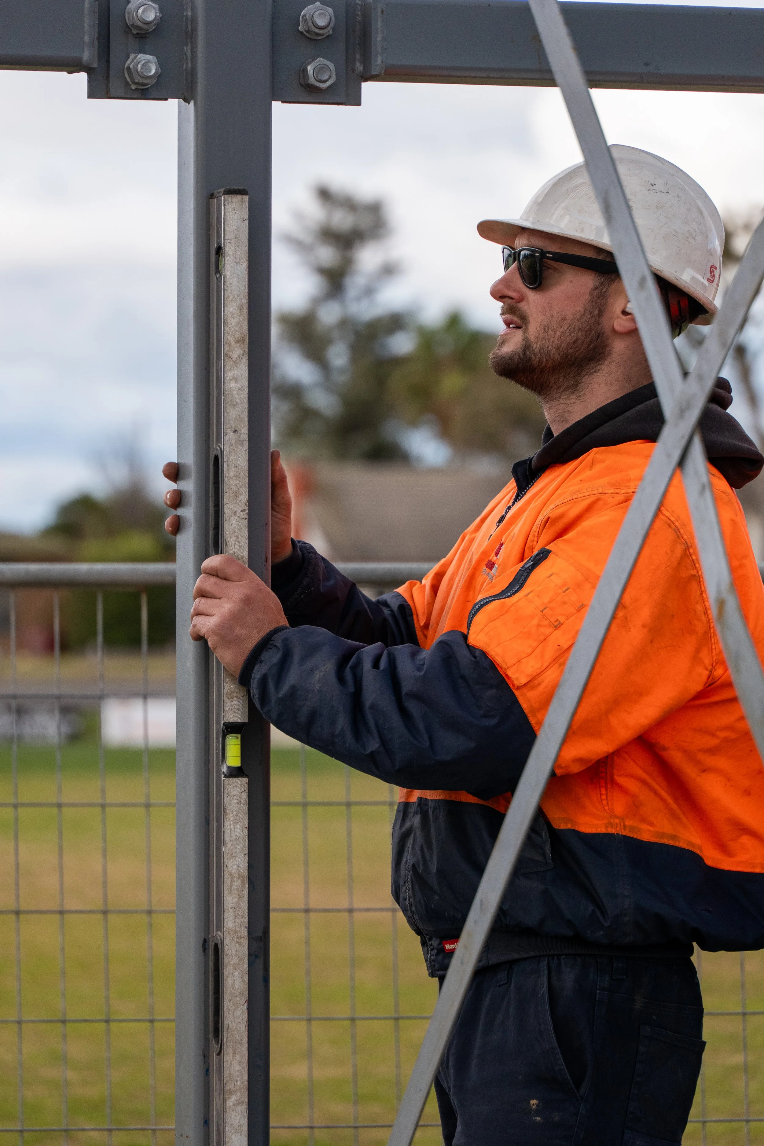 A construction worker wearing a white hard hat, sunglasses, and an orange safety vest working on a metal structure, with a fence and cloudy sky in the background.