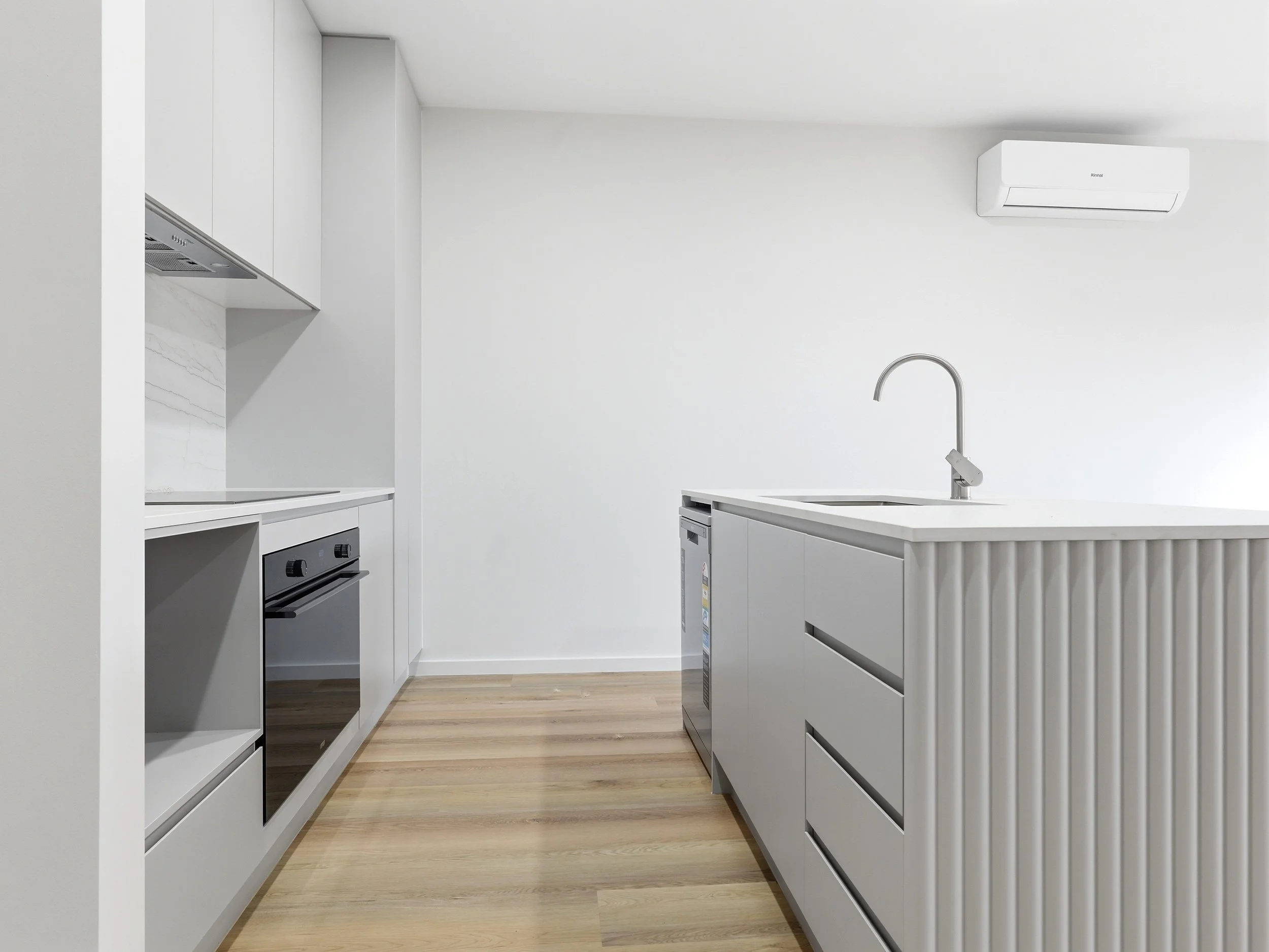 Modern white kitchen with light wood flooring, white cabinets, an oven, a sink with a curved faucet, and wall-mounted air conditioning unit.