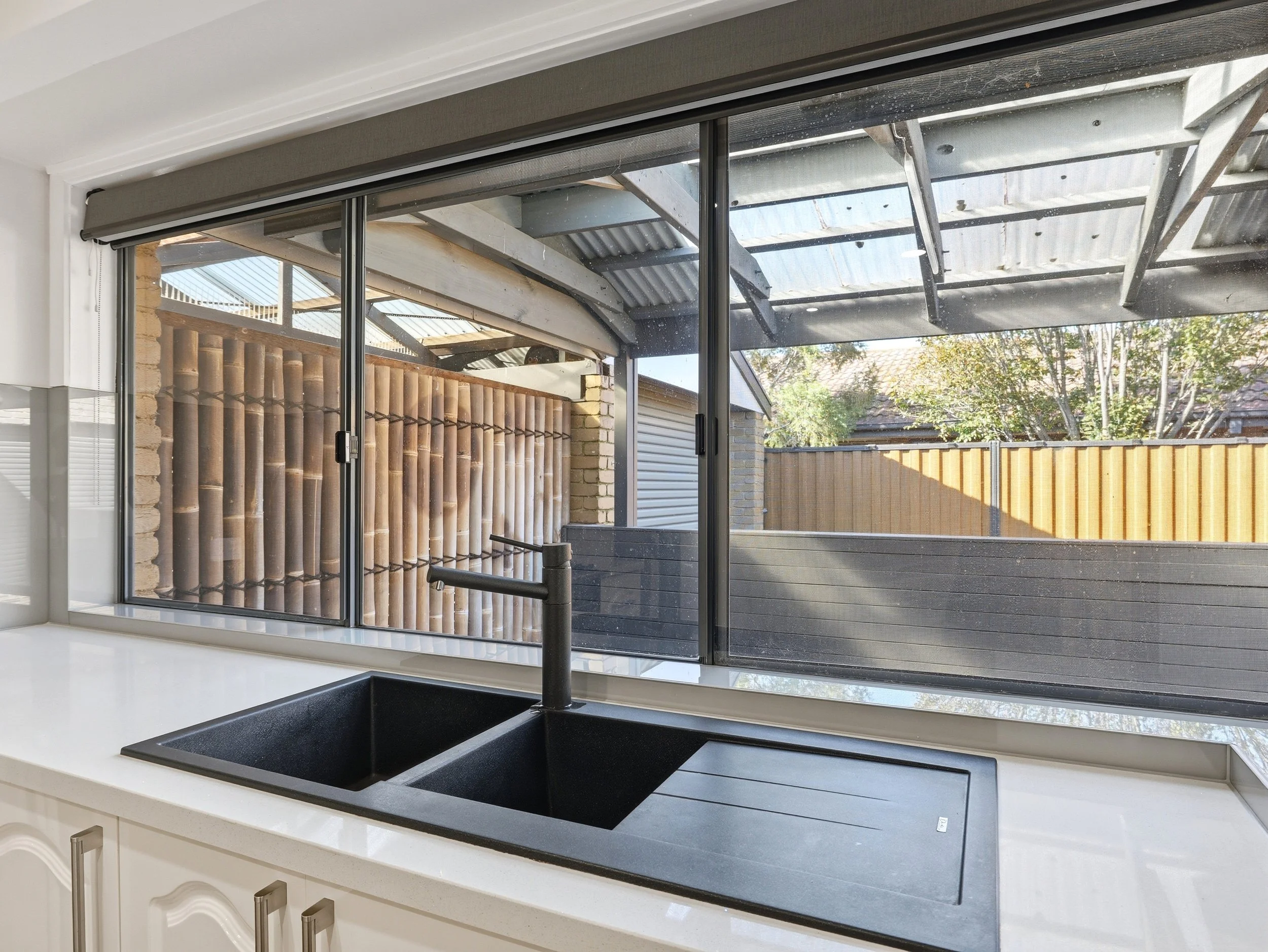 View from inside a kitchen looking out through a large glass sliding door. The kitchen has a white countertop with a black sink and a black faucet. The outside area shows a wooden fence, trees, and part of a roof.
