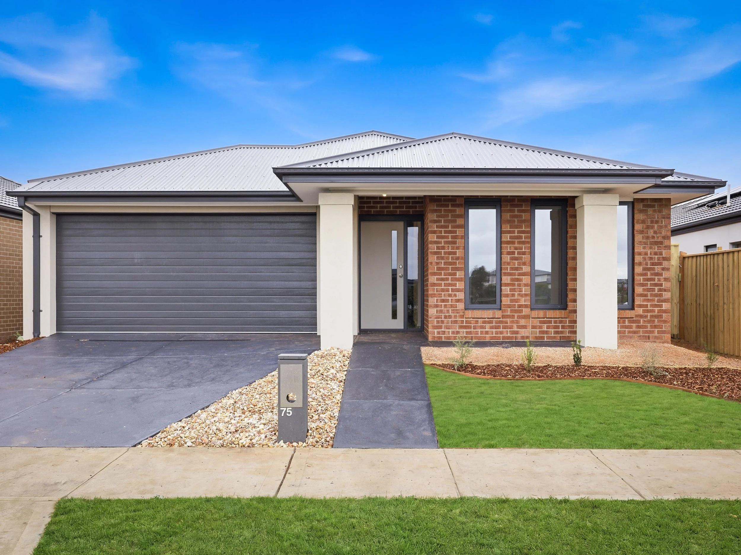 A modern house with a grey garage door, brick and cream exterior, and a front yard with a concrete walkway, gravel, and grass. The house has a metal roof and a small porch with a glass door, under a clear blue sky.