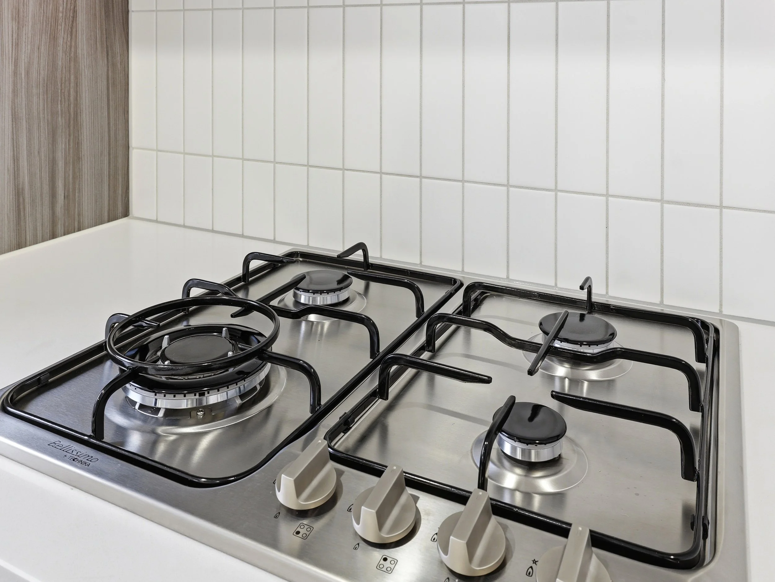 A white gas stove with four burners and black grates, installed on a white countertop in a kitchen with white tiled wall backsplash.