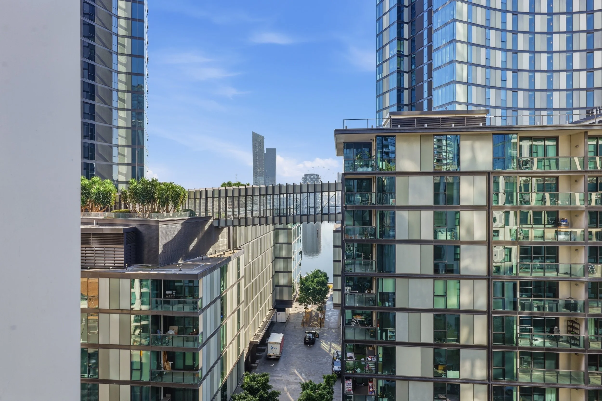 View of modern glass buildings with a water canal in between, greenery on top of the buildings, and a clear blue sky in the background.
