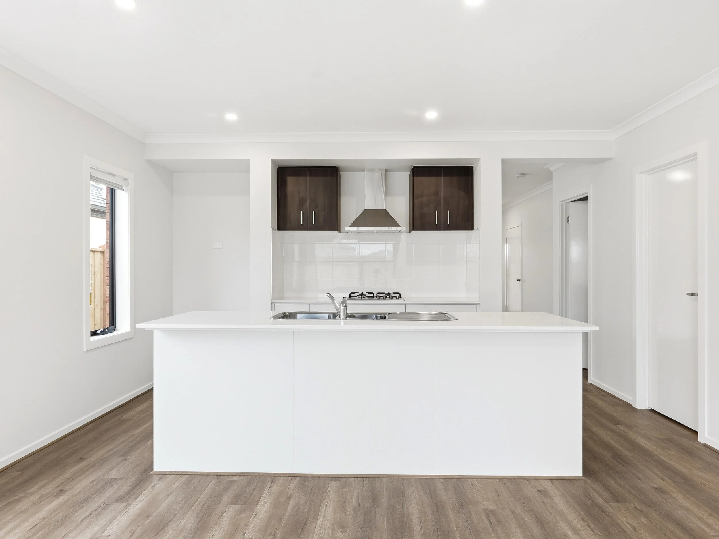Modern kitchen with white island counter, dark upper cabinets, stainless steel range hood, white tiled backsplash, two windows, and wood flooring.