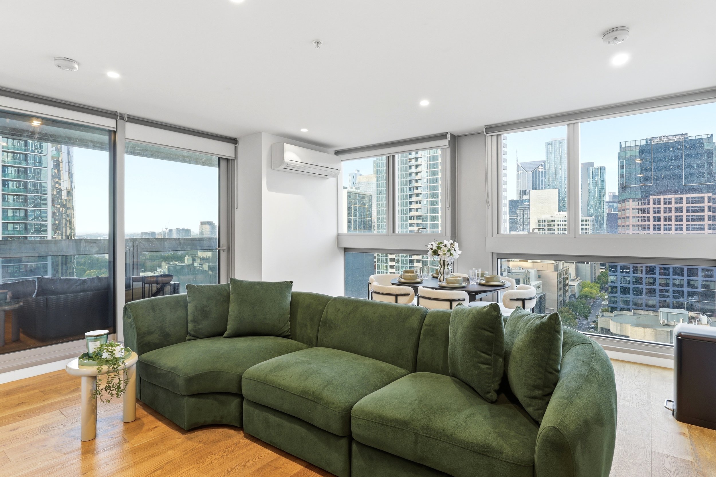 Interior of a modern apartment with large windows providing a city view, green sectional sofa, round table with dishes, and a small side table with a plant.