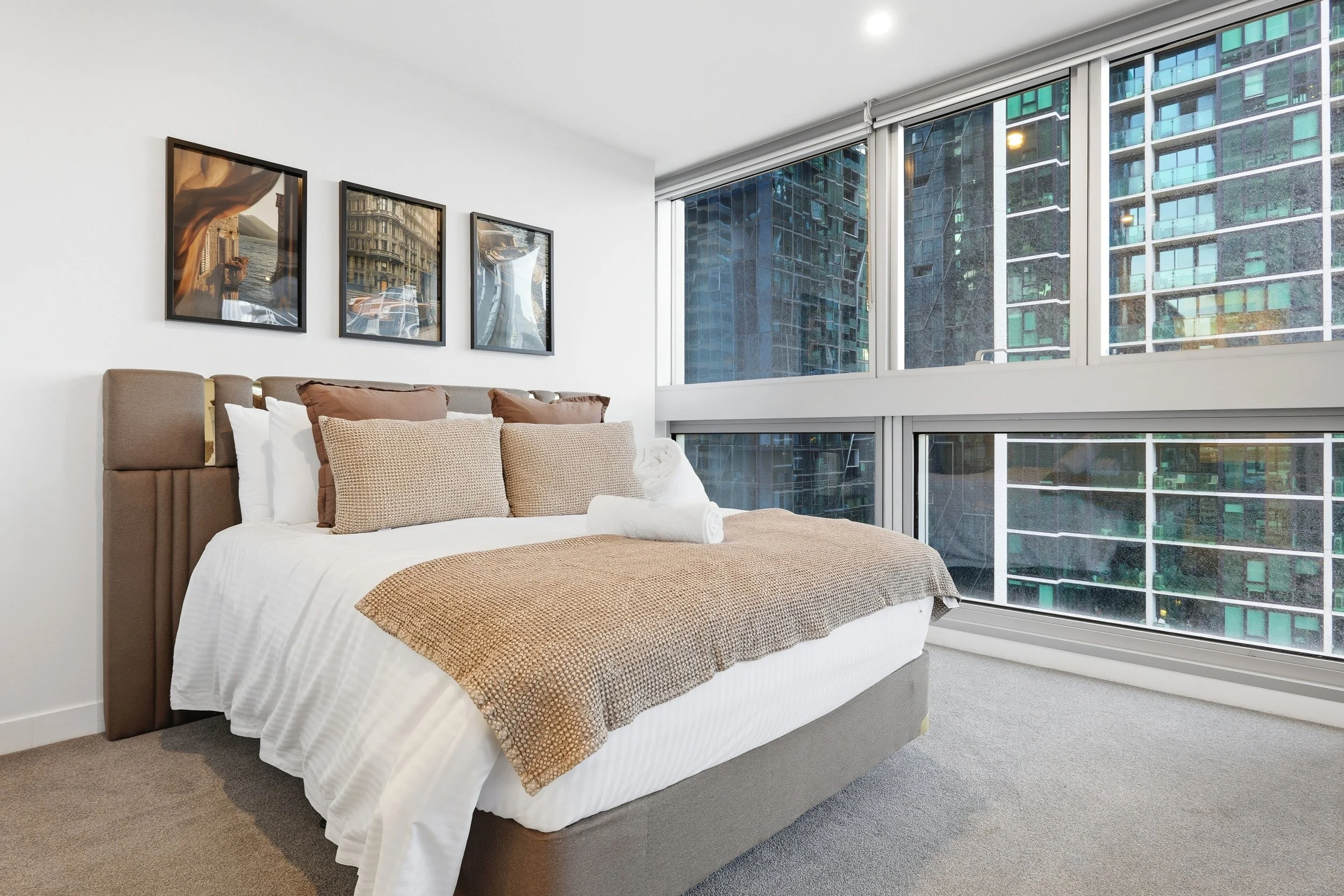 Modern bedroom with a neatly made bed featuring white linens, brown pillows, and a tan blanket. The room has large floor-to-ceiling windows with a view of neighboring high-rise buildings.