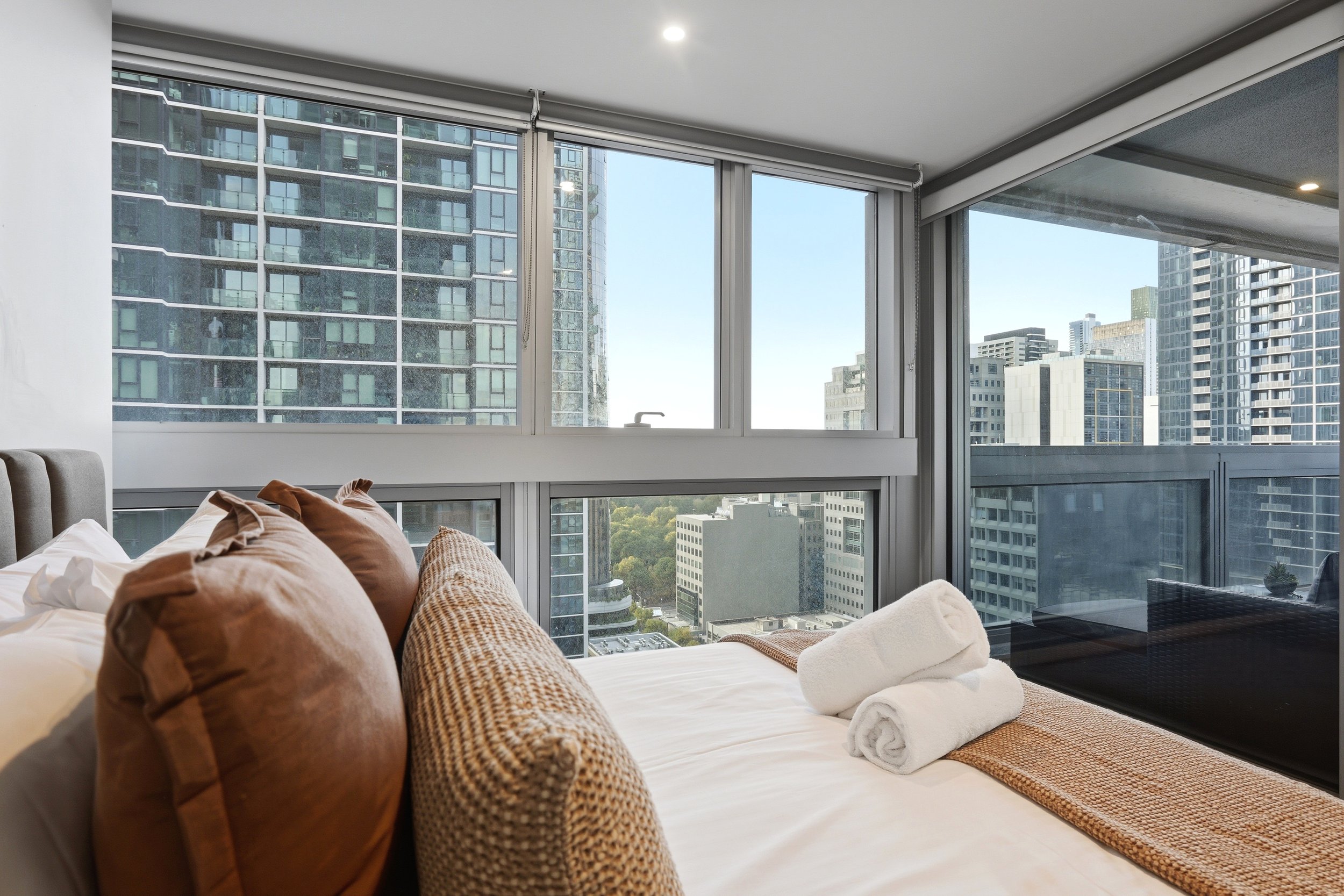 Interior view of a modern apartment bedroom with large windows showing skyscrapers outside, a bed with brown and white pillows, a rolled towel, and a woven rug.