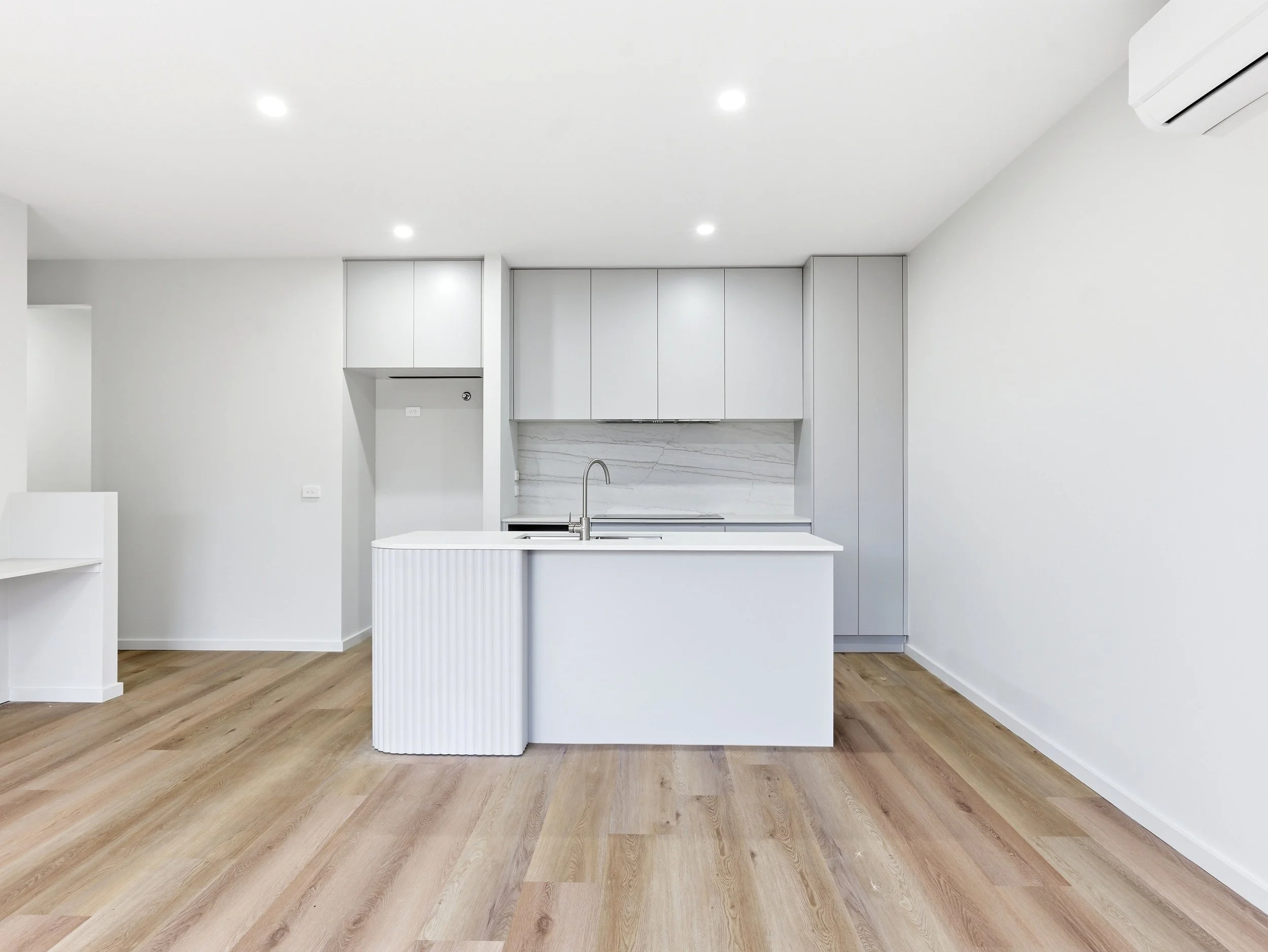 A modern, minimalist kitchen with a white island, cabinetry, and marble backsplash. The floor is wooden, and there are ceiling lights and an air conditioning unit.