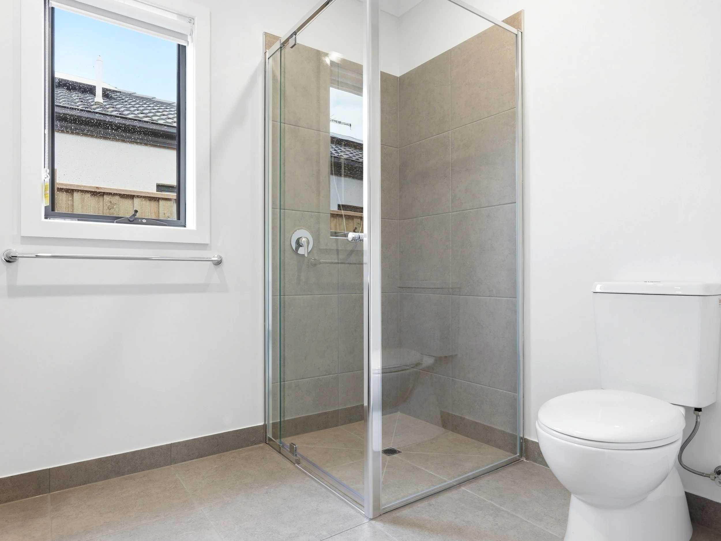 A modern bathroom with a toilet, a glass shower enclosure, a small window with a view of a house roof outside, beige tiled shower walls, white painted walls, and a towel bar next to the window.