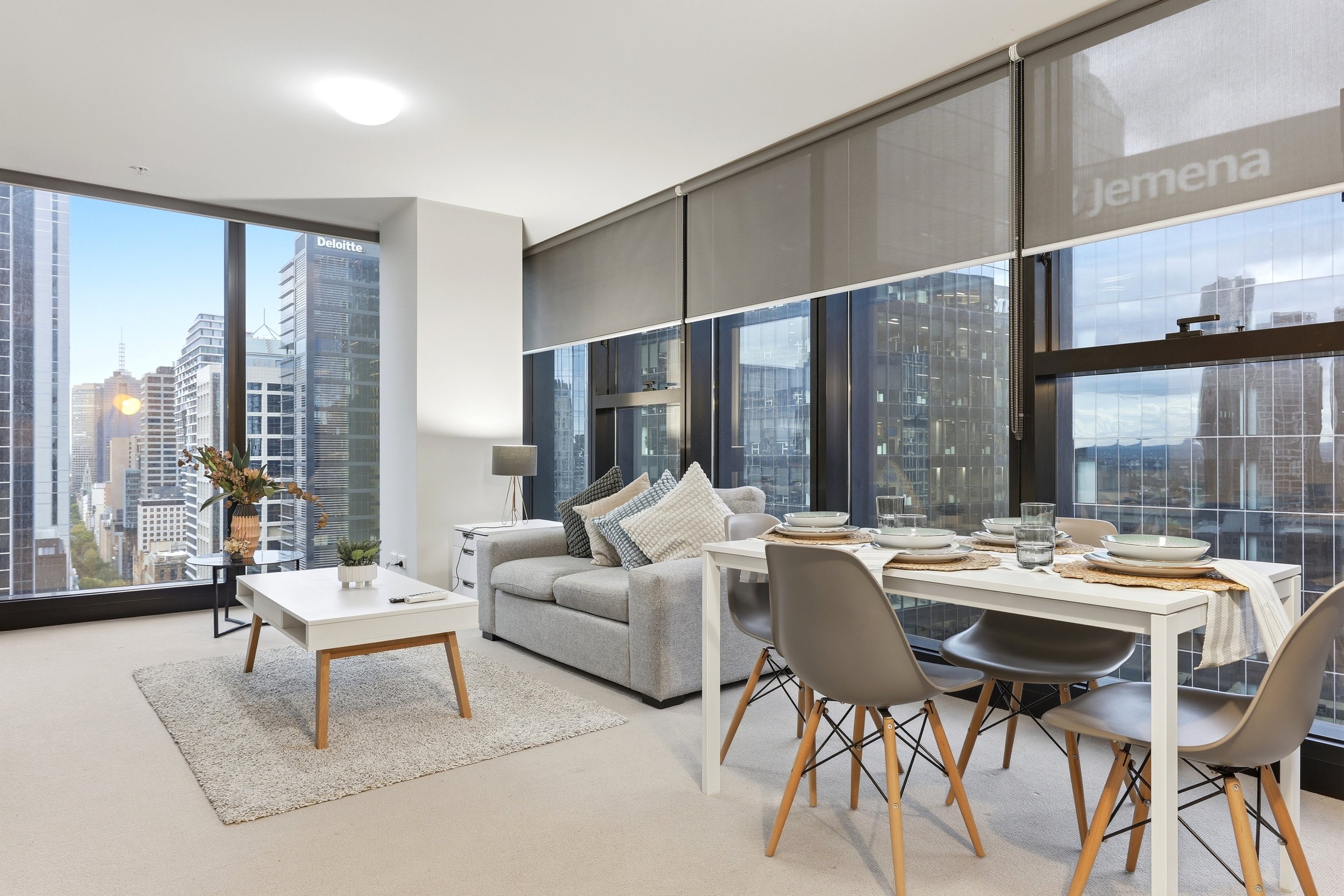 Modern high-rise apartment living room with large windows, city skyline view, grey sofa with pillows, coffee table, dining table with plates and glasses, and minimalist decor.