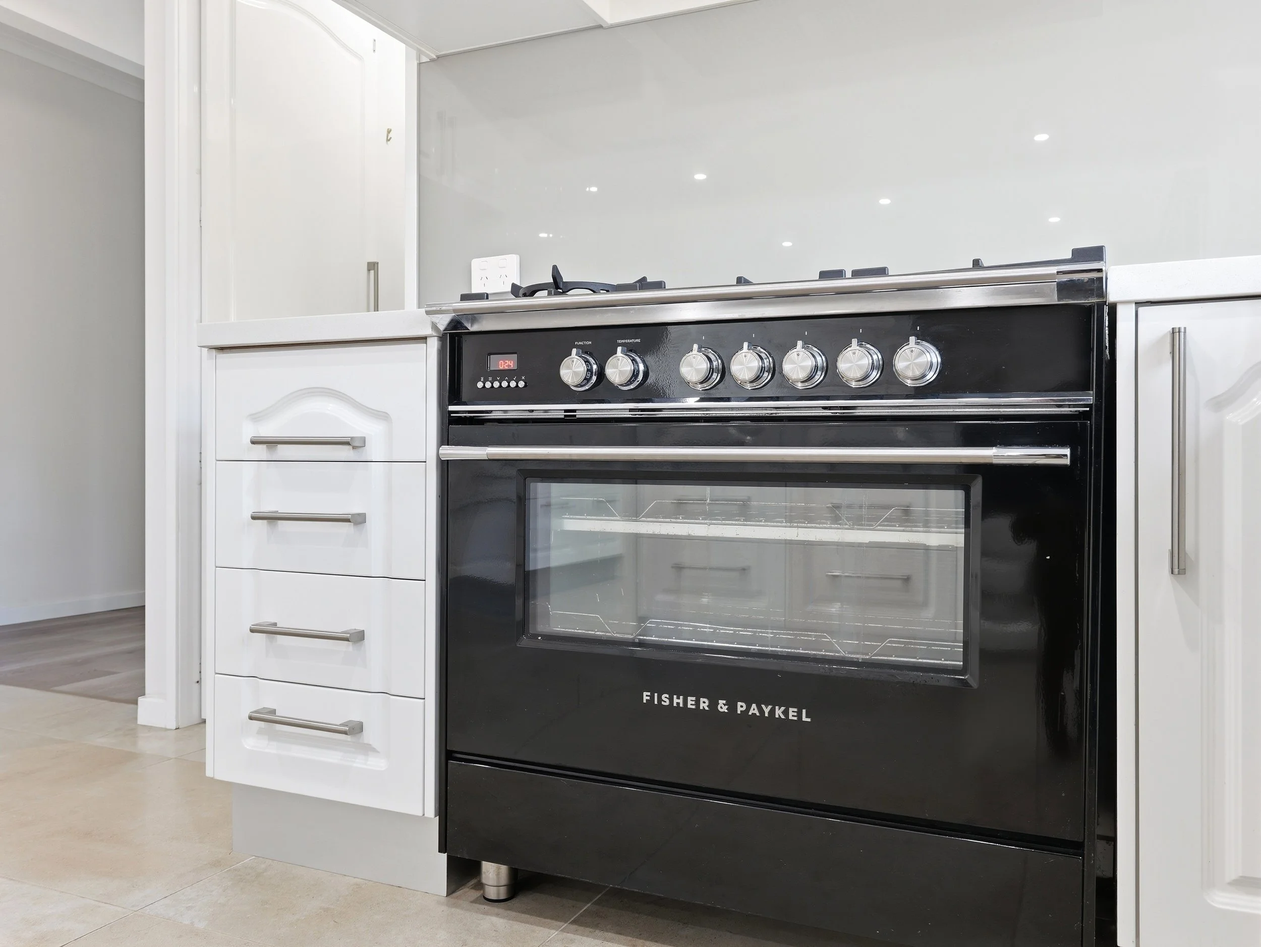 Black and silver Fisher & Paykel oven with knobs, built into white kitchen cabinets, in a modern kitchen with beige tile floor.
