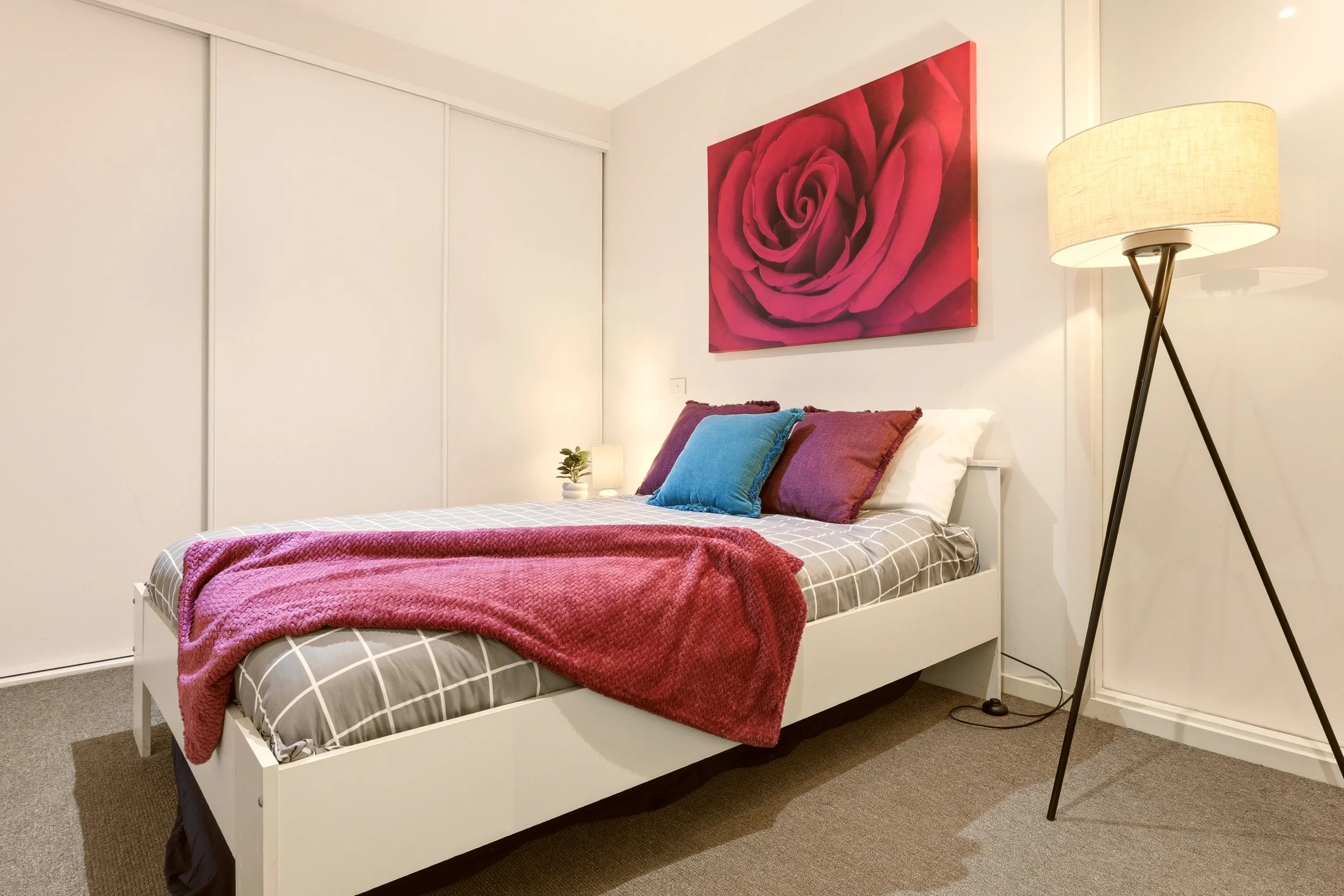 A bedroom with a white bed frame and checkered bedding in gray and white. The bed has multiple pillows in shades of blue, purple, and red. A large red rose flower painting hangs on the wall above the bed. To the right, there is a floor lamp with a be