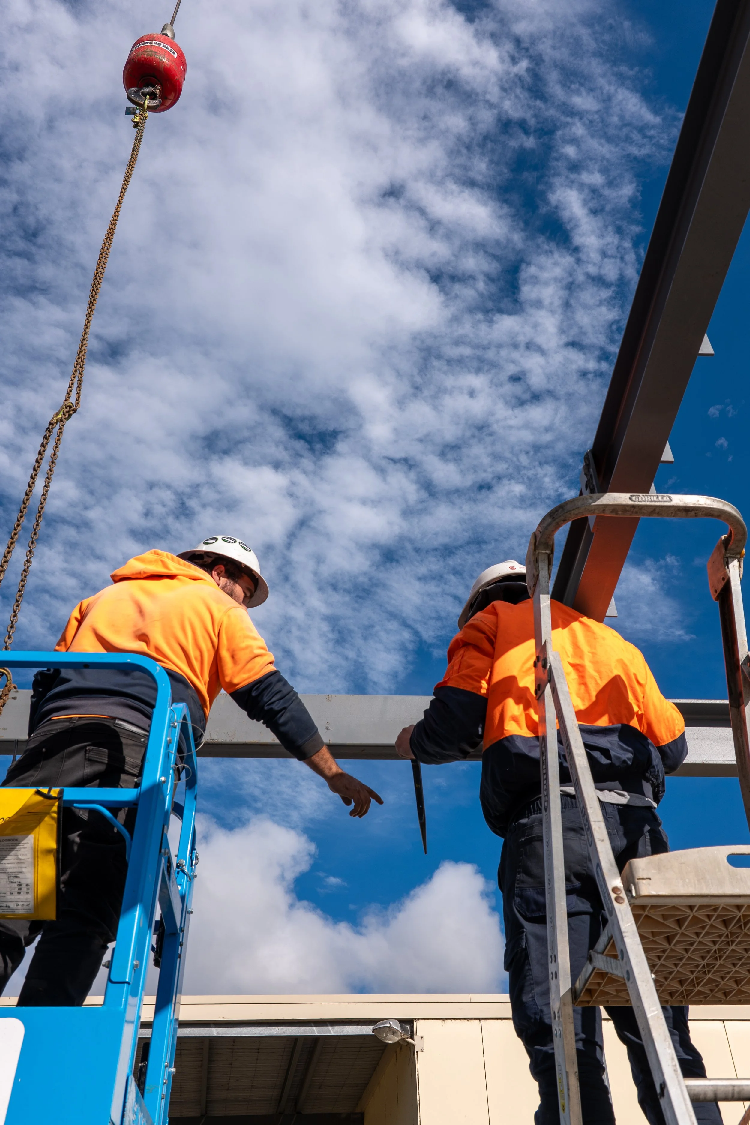 Two construction workers, wearing orange jackets and helmets, are working on a construction site. They are standing on a scaffold, working on a metal beam with a crane overhead, under a partly cloudy sky.