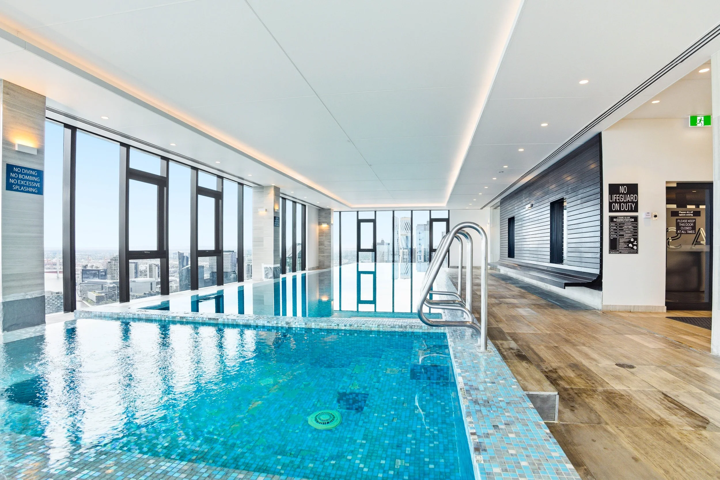 Indoor rooftop pool with large windows overlooking city skyline, stainless steel ladder, tiled floor, and modern lighting.