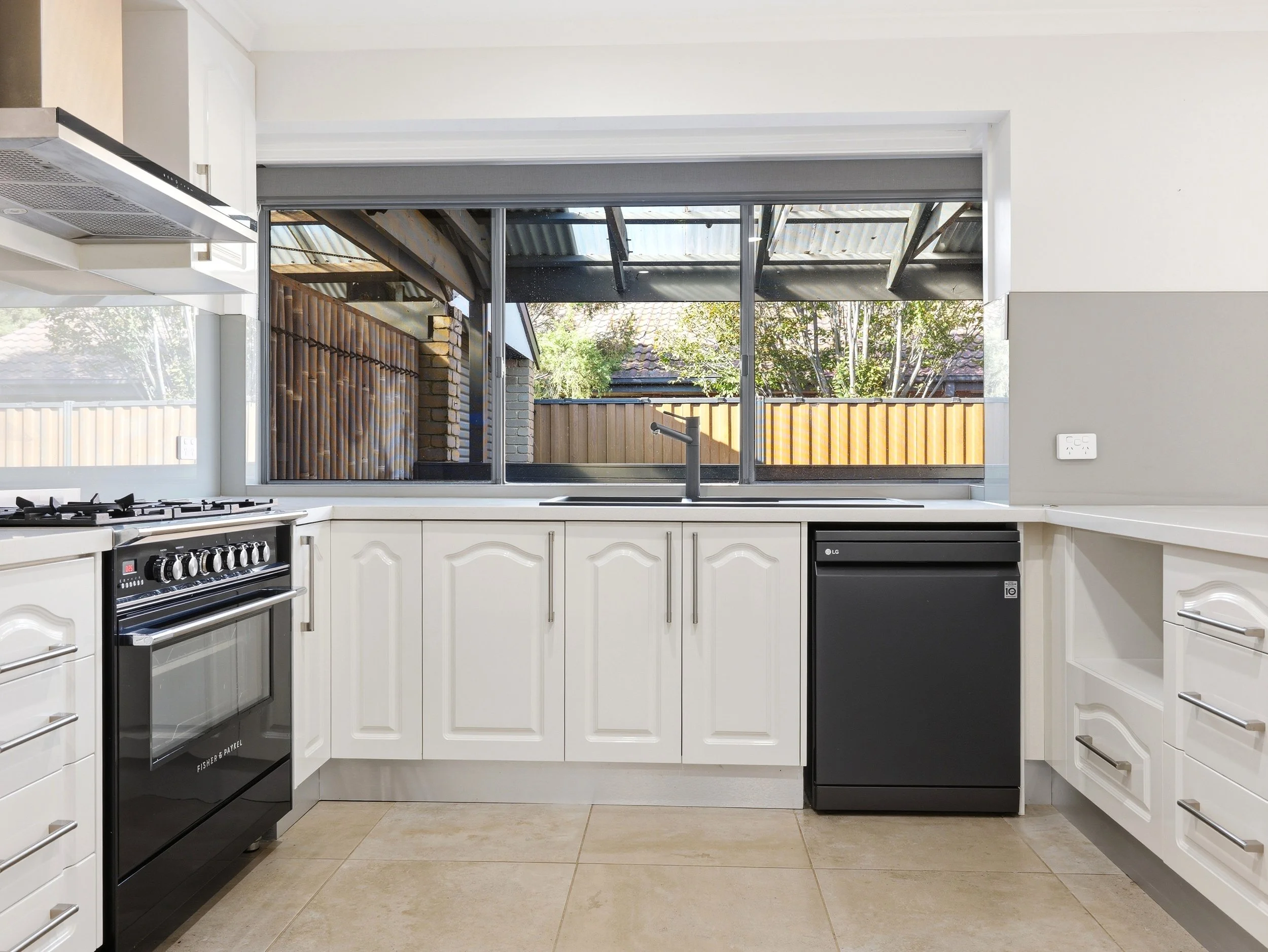 Kitchen with white cabinets, black oven, and large window showing backyard with trees and brick wall.