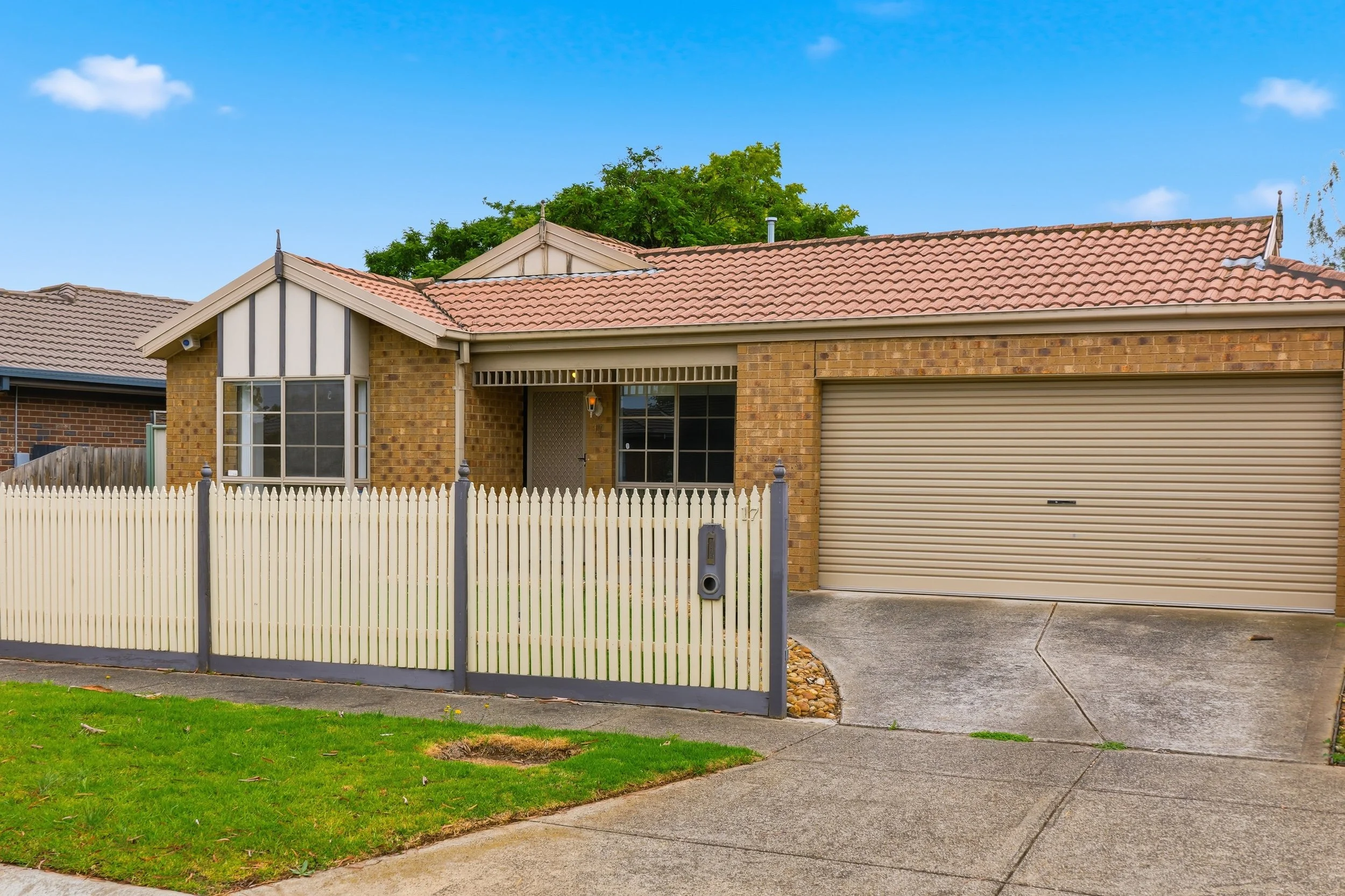 Front view of a brick house with a red tile roof, white picket fence, and a beige roller garage door. The house has two windows and a small front lawn with a concrete driveway and pathway.