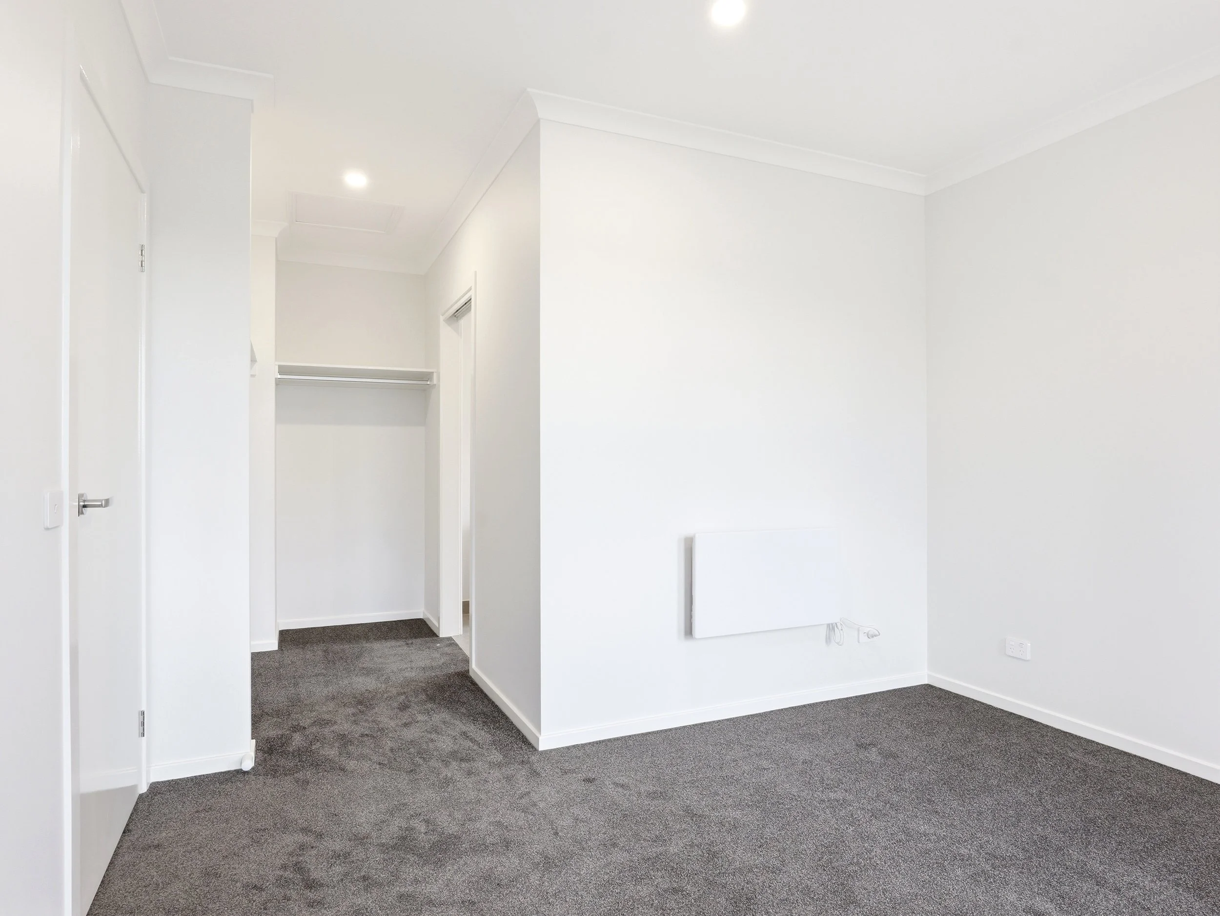 Empty room with white walls and gray carpeted floor, including a door on the left and a wall-mounted radiator.