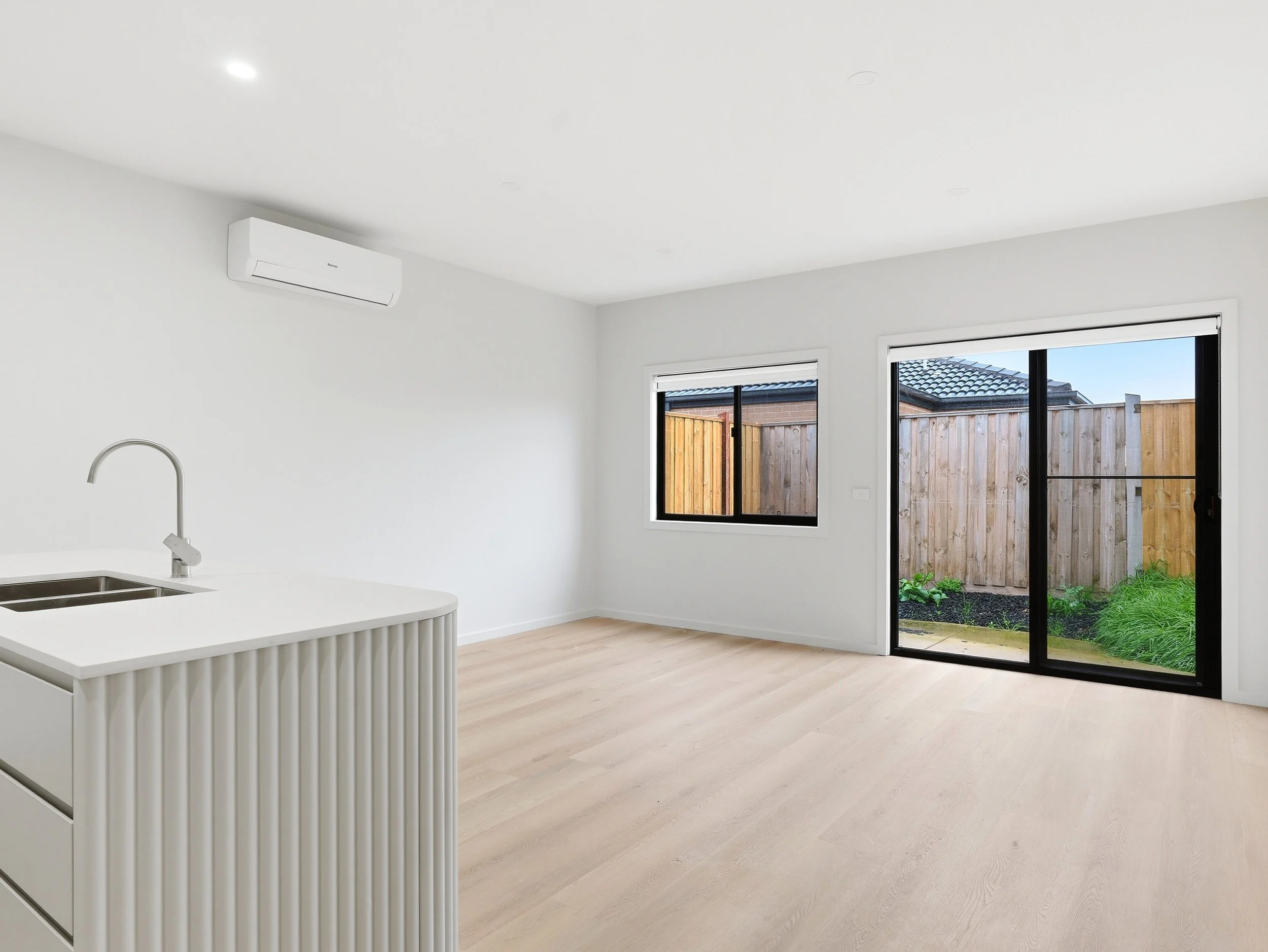 The image shows a bright, modern kitchen and dining area with light wood flooring, white walls, and large windows. There is a sliding glass door leading to an outdoor fenced yard, and an air conditioning unit is mounted on the wall.