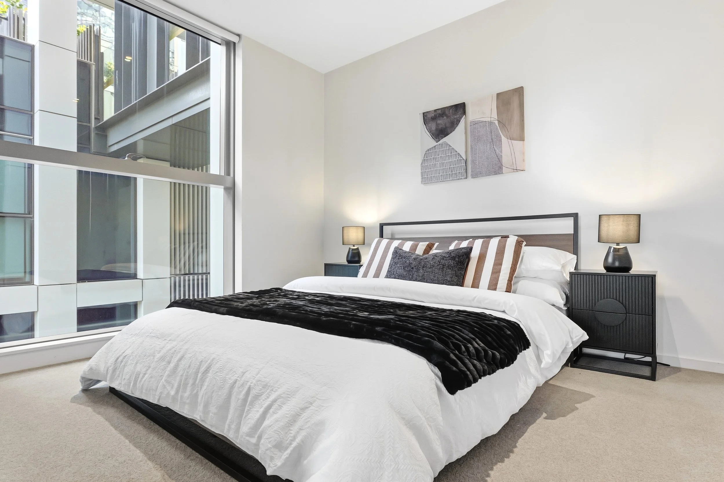 A modern bedroom with a large bed featuring white and striped bedding, black velvet throw, and dark pillows. There are two black nightstands with matching lamps on each side of the bed. A window on the left side brings in natural light. Abstract wall