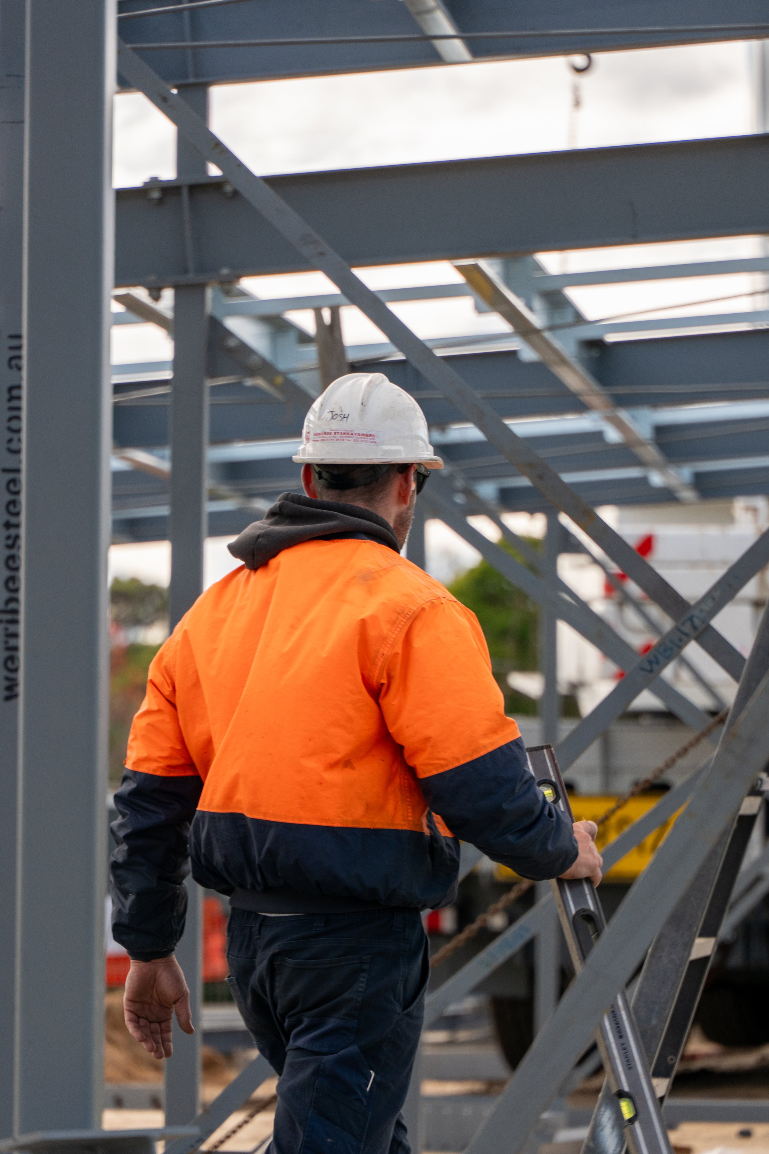 A construction worker wearing an orange safety jacket, a white helmet with the name 'Josh' written on it, and safety glasses working on a steel framework structure at a construction site.