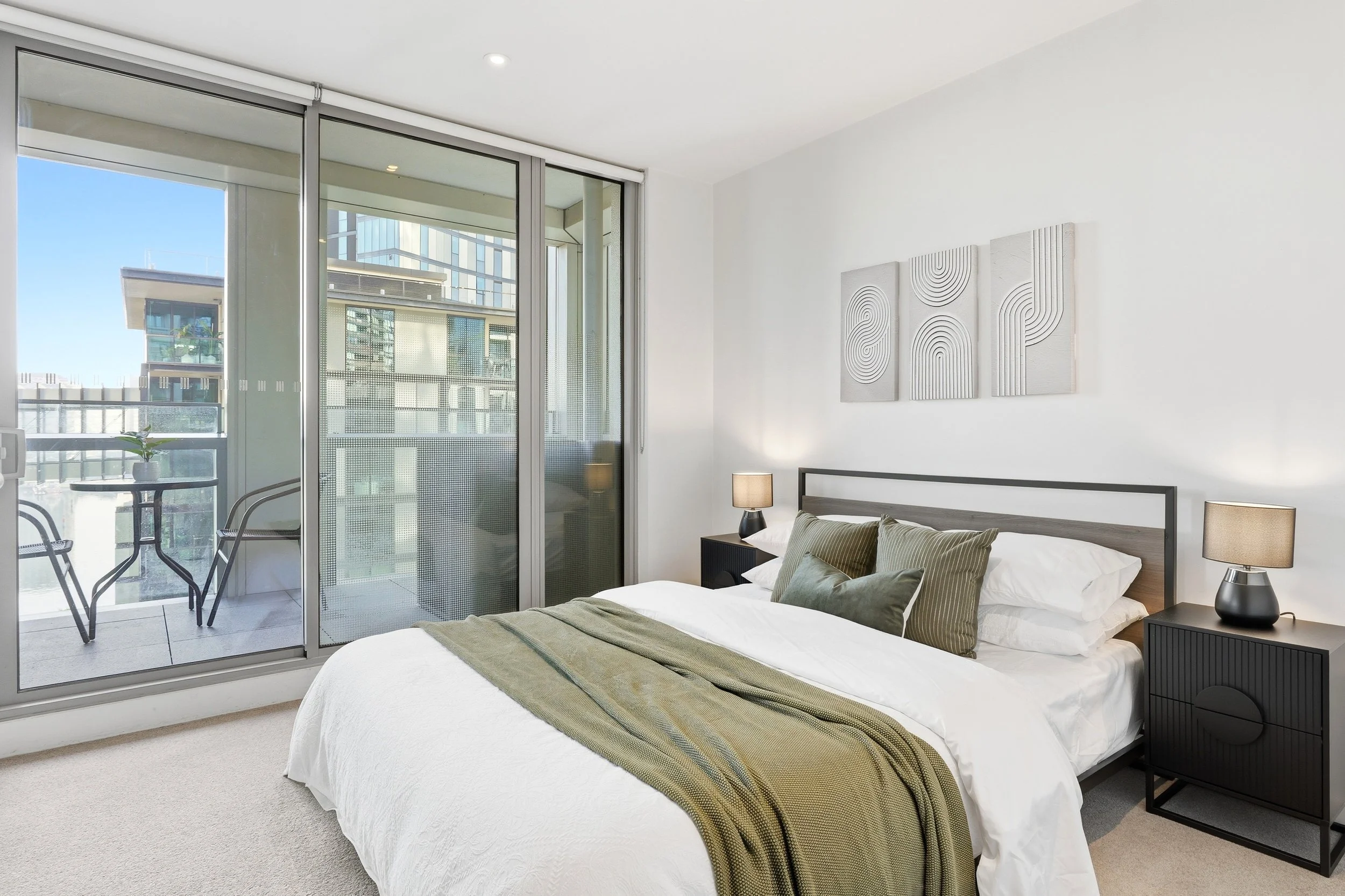 Modern bedroom with a balcony, featuring a large bed with white bedding and green accents, black bedside tables with lamps, wall art, and a sliding glass door leading to the balcony with outdoor chairs and a small table.