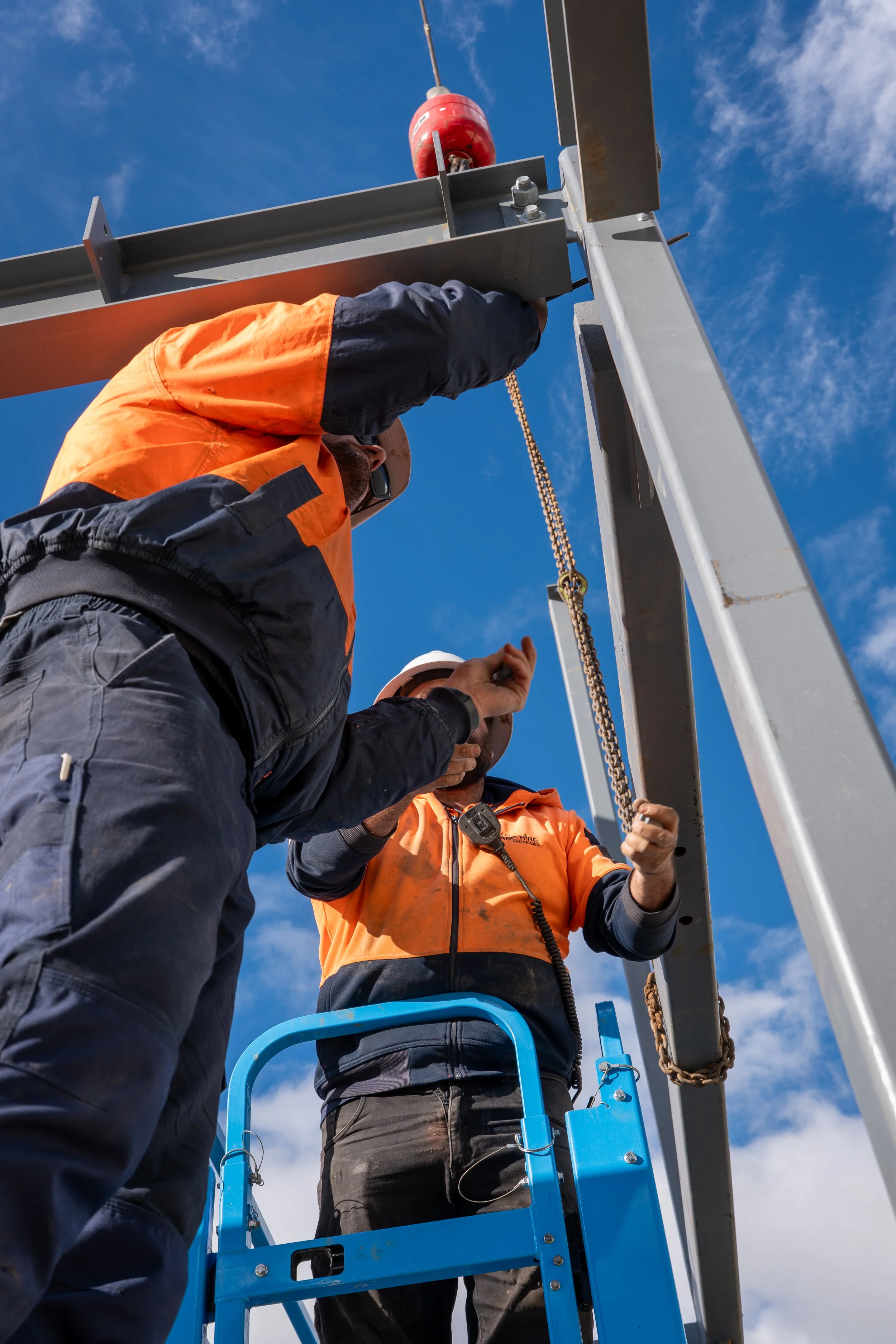 Workers in orange safety vests installing or maintaining a tall metal structure outdoors, with blue sky and clouds in the background.