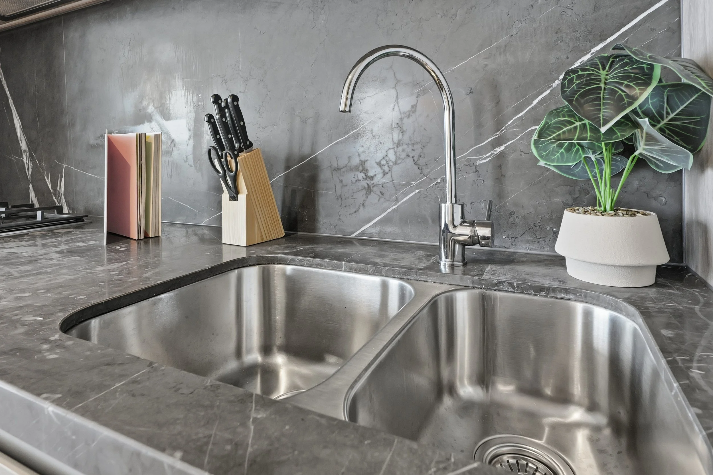 Kitchen sink with a gray marble countertop, a modern chrome faucet, a plant in a white pot, a knife block, and a set of knives on the countertop.