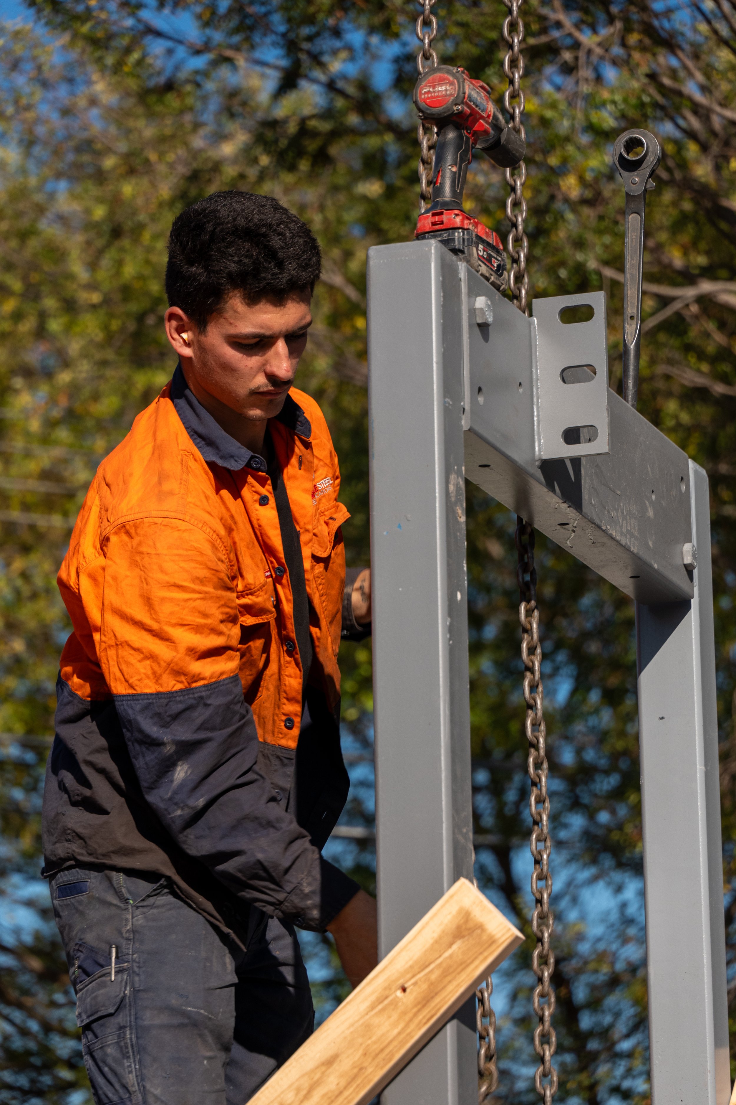 A worker in an orange and gray jacket is working on a metal structure with a chain and a cordless drill hanging from it, outdoors with trees in the background.