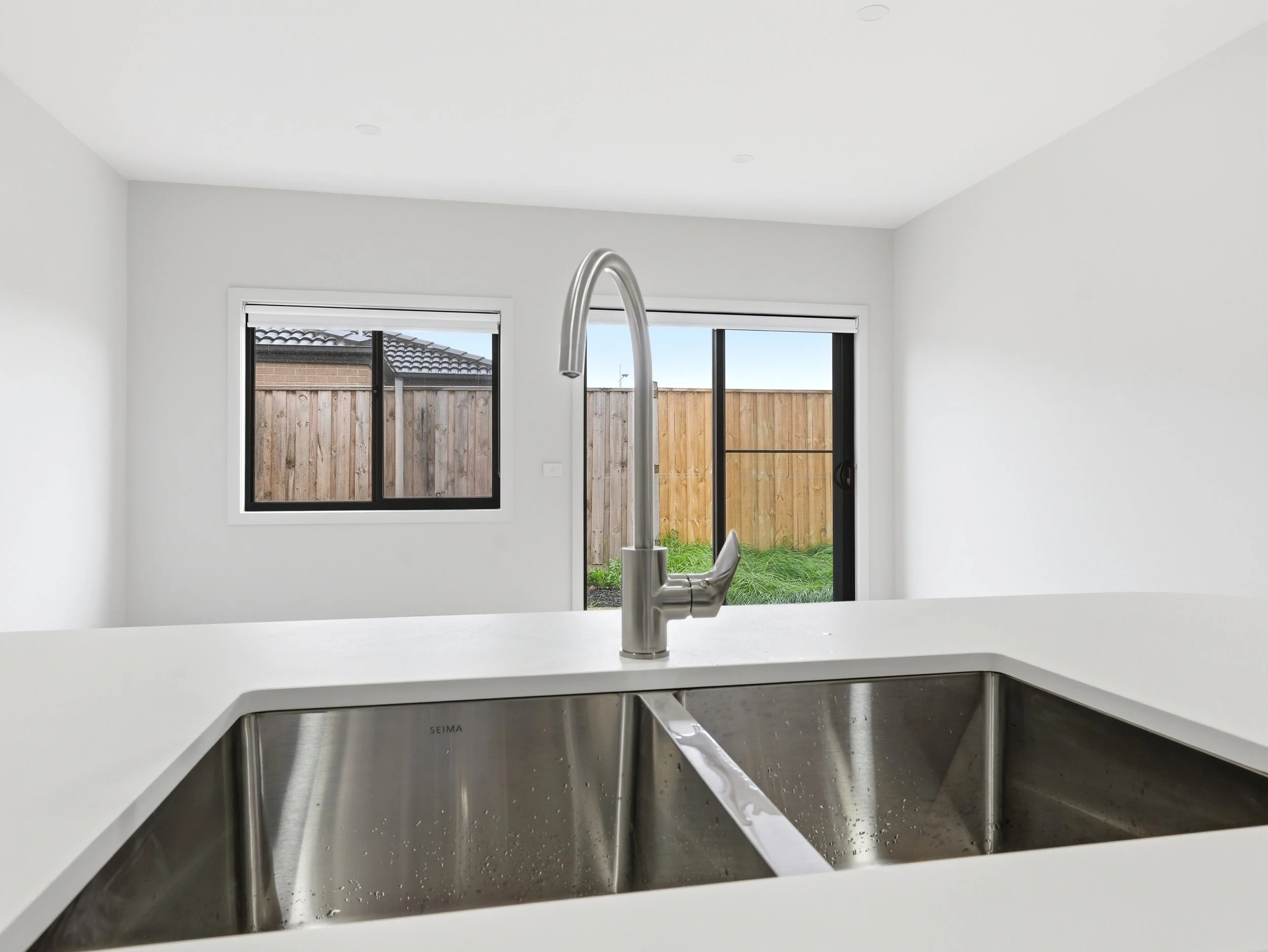 Modern kitchen with white walls, stainless steel sink, and large windows showing a backyard with a wooden fence and neighboring house roof.