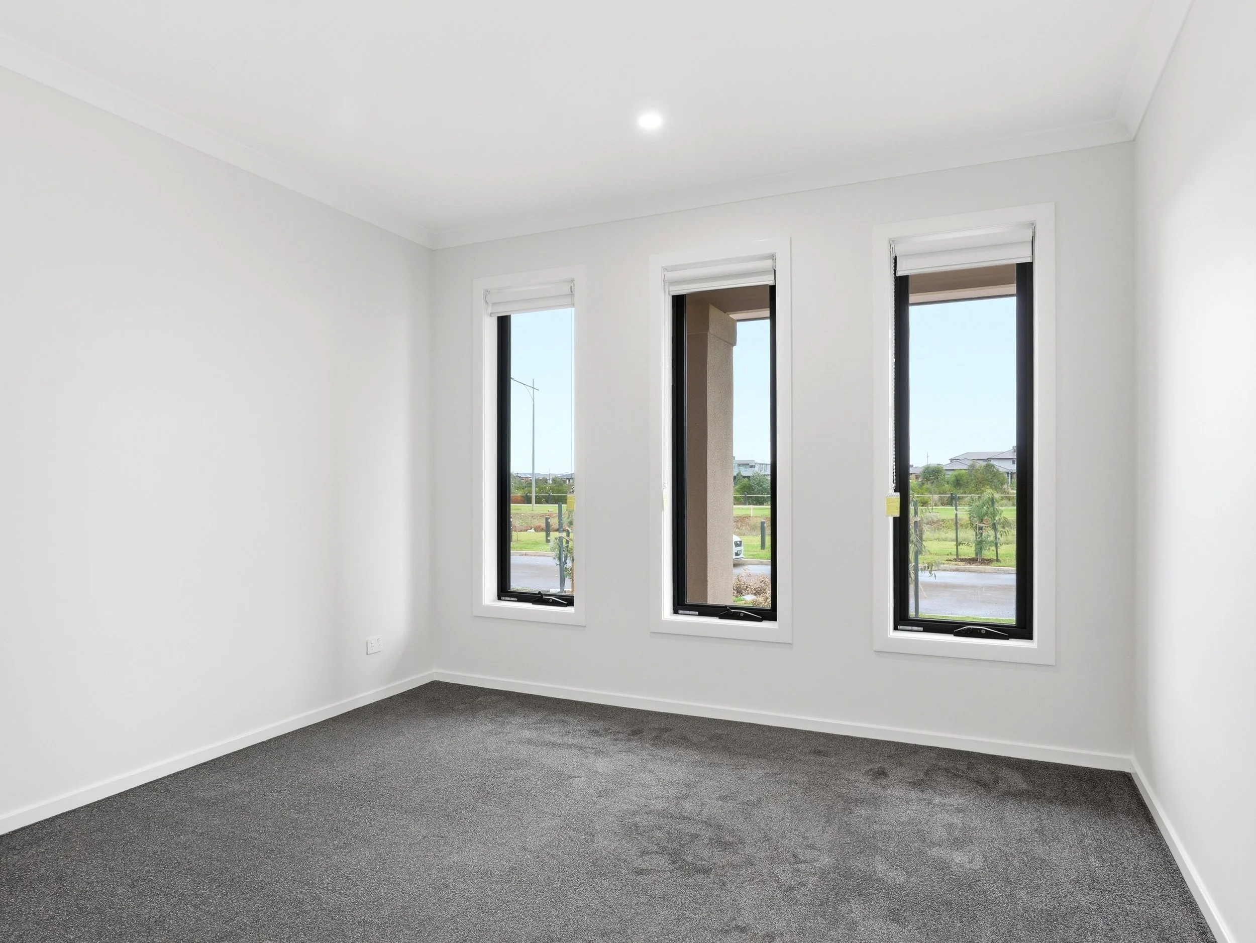 Empty room with white walls, a dark gray carpet, and three tall windows showing a view outside of houses, trees, and a cloudy sky.
