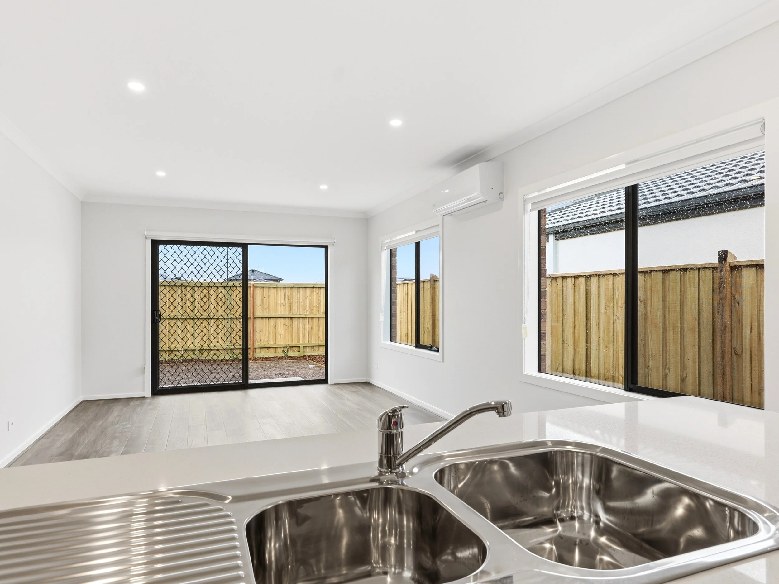 View from kitchen sink looking into a bright living room with large windows, sliding door, and outdoor fenced area.