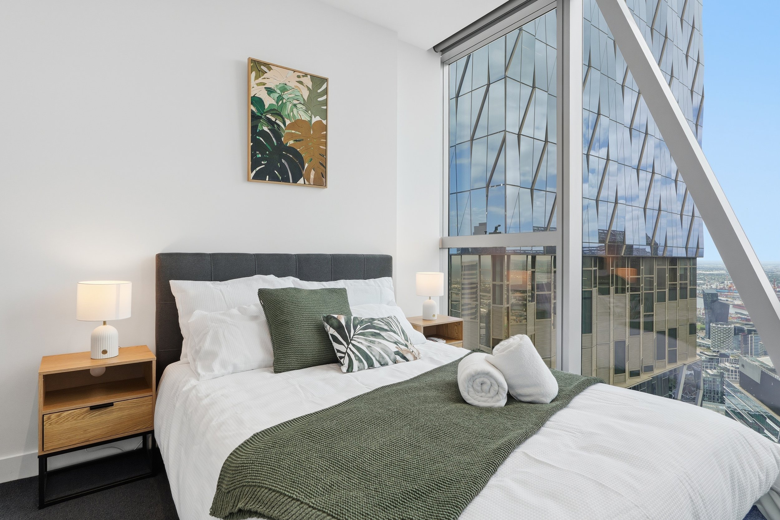 Modern bedroom with a large window overlooking a city, a bed with white bedding, green pillows, and a green blanket, two rolled towels, nightstands with white lamps, and a piece of art featuring tropical leaves on the wall.