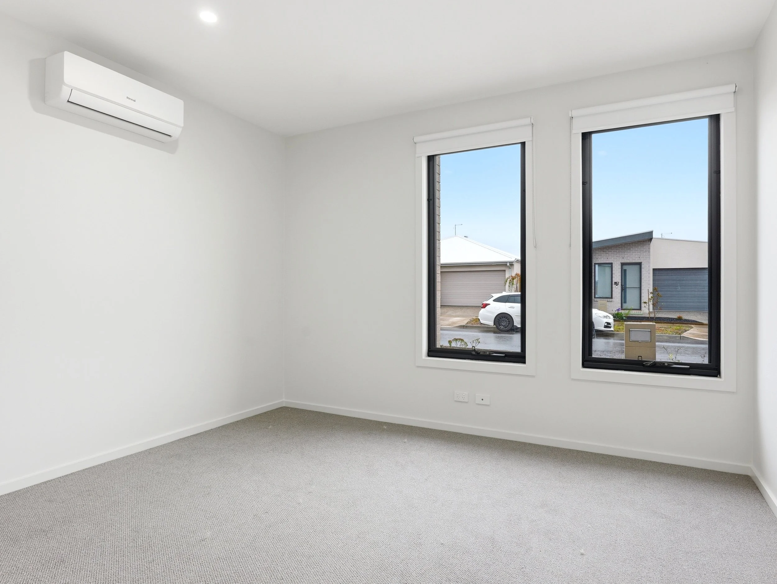 Empty room with white walls, gray carpet, and large windows showing a residential neighborhood outside.