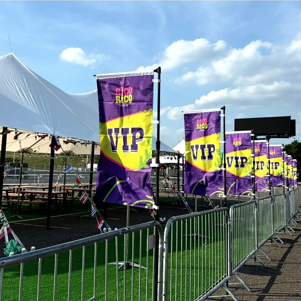 Event area with purple VIP flags, picnic tables, and a large white tent under a partly cloudy sky.