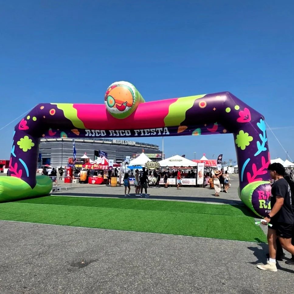 Inflatable archway with colorful floral and graphic designs at an outdoor festival, with tents and people in the background and clear blue sky.