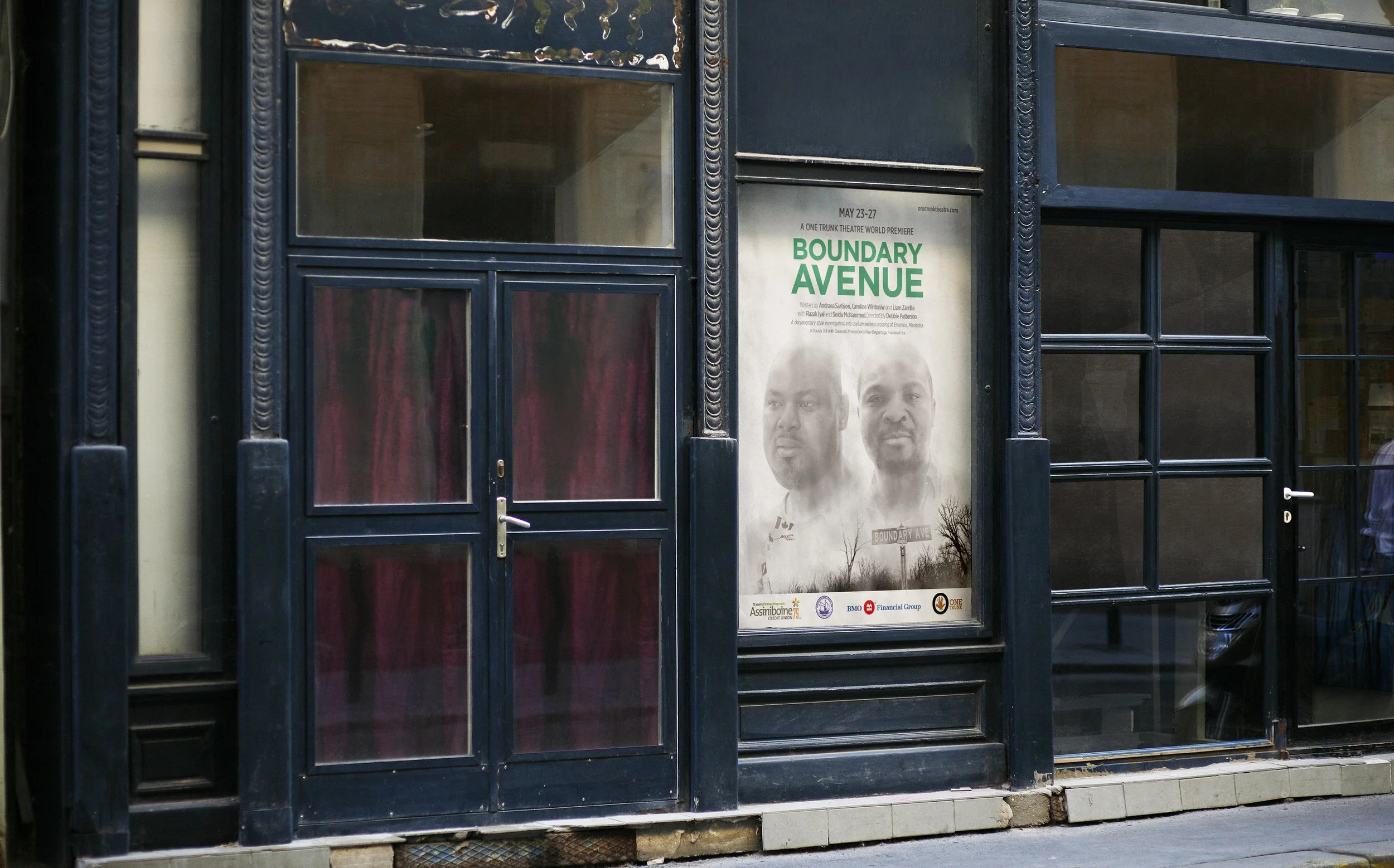A poster for the production of 'Boundary Avenue' inside a glass-fronted storefront, with two men's faces and text about the premiere event.