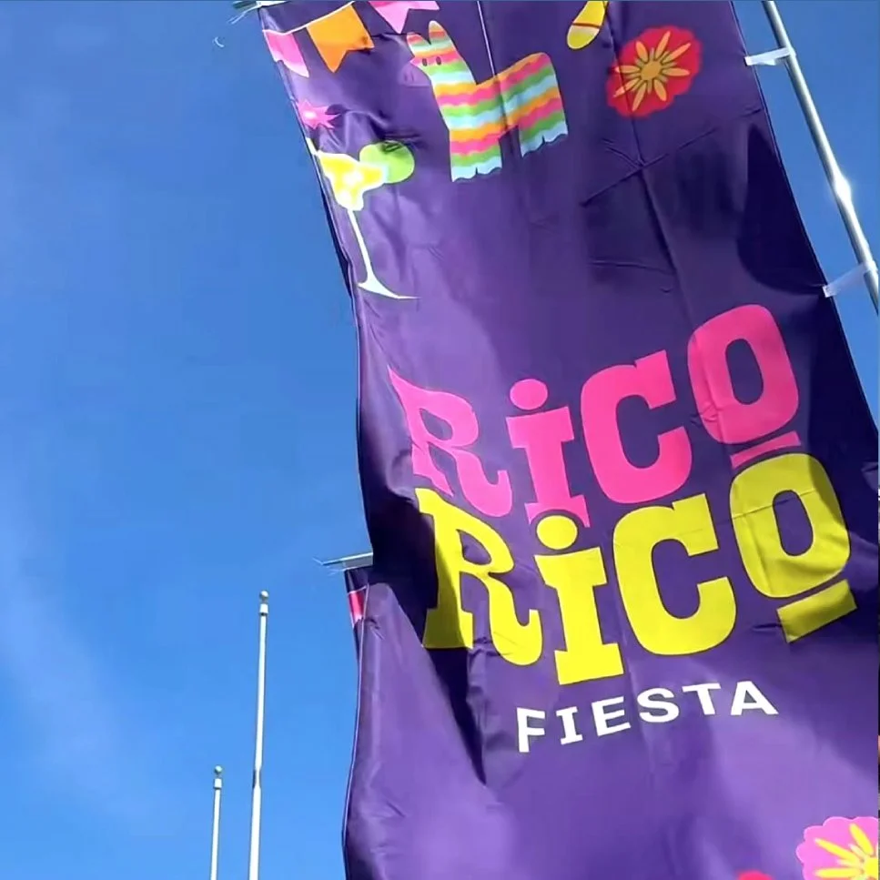 Colorful flags with the words "Rico Rico Fiesta" on a purple background, flying outdoors against a clear blue sky.