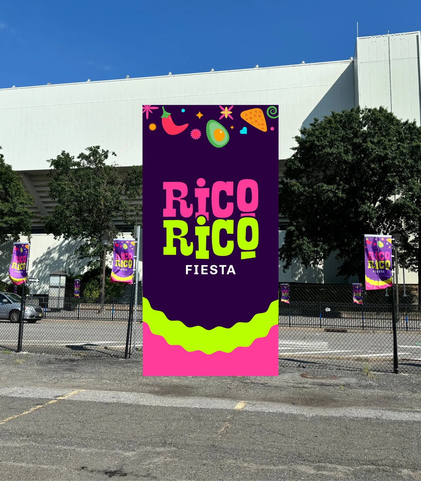 A large vertical sign in front of a building advertising Rico Rico Fiesta, with colorful fruits and food illustrations at the top, including a chili pepper, avocado, strawberry, and tortilla chip, set against a dark purple background.