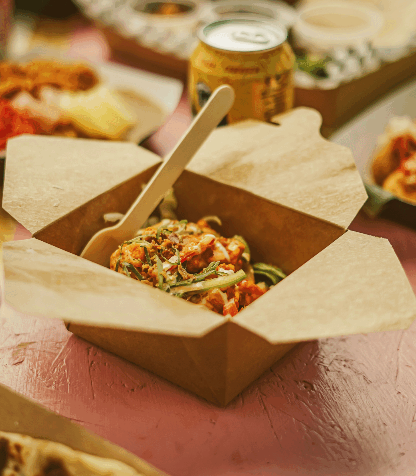 Takeout box of spicy chicken and vegetables with herbs and spices, on a pink wooden table, with a can of soda and other food in the background.