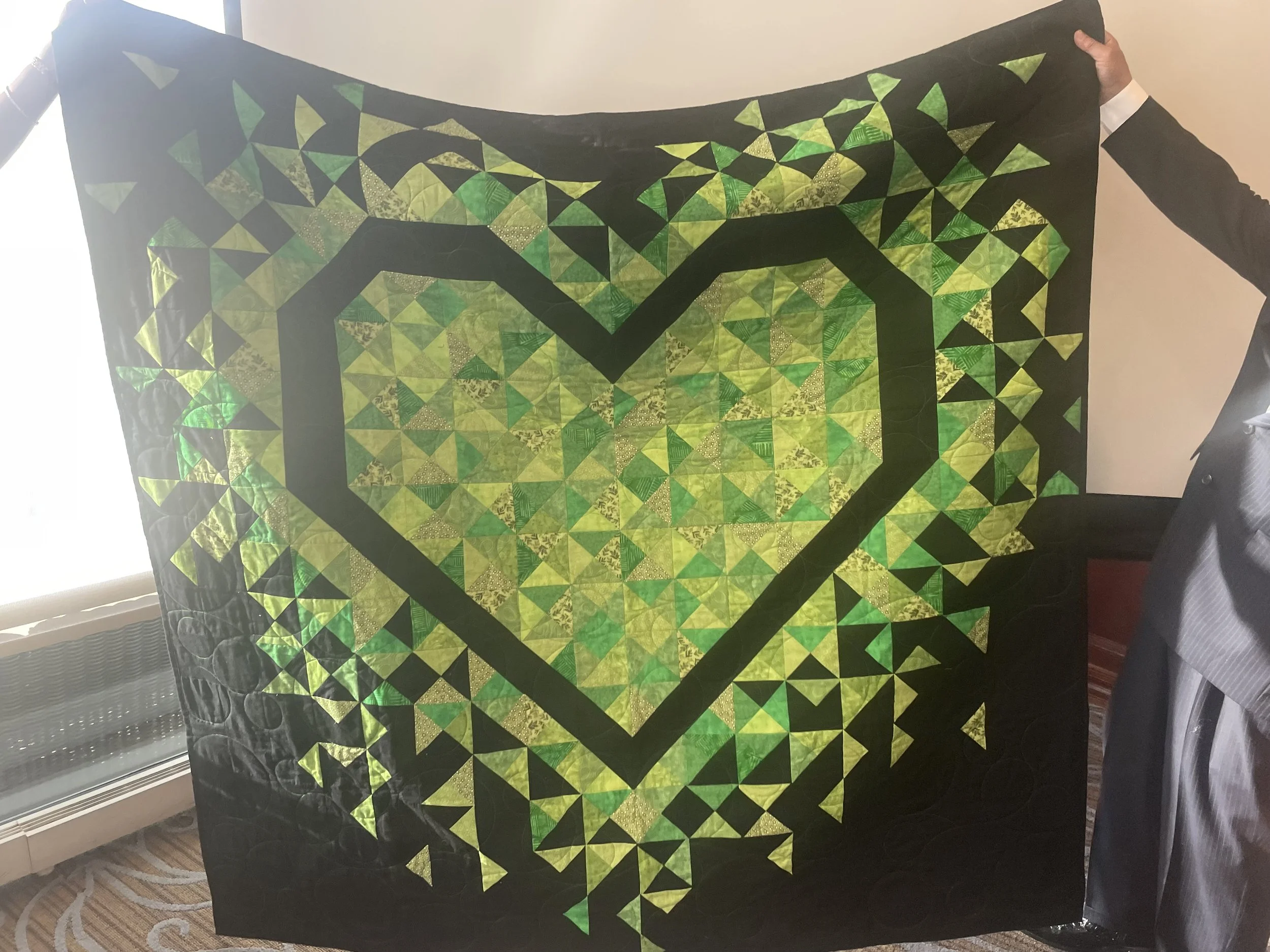 Two people hold up a quilt with a large heart design in the center, made of green and yellow triangles on a black background.
