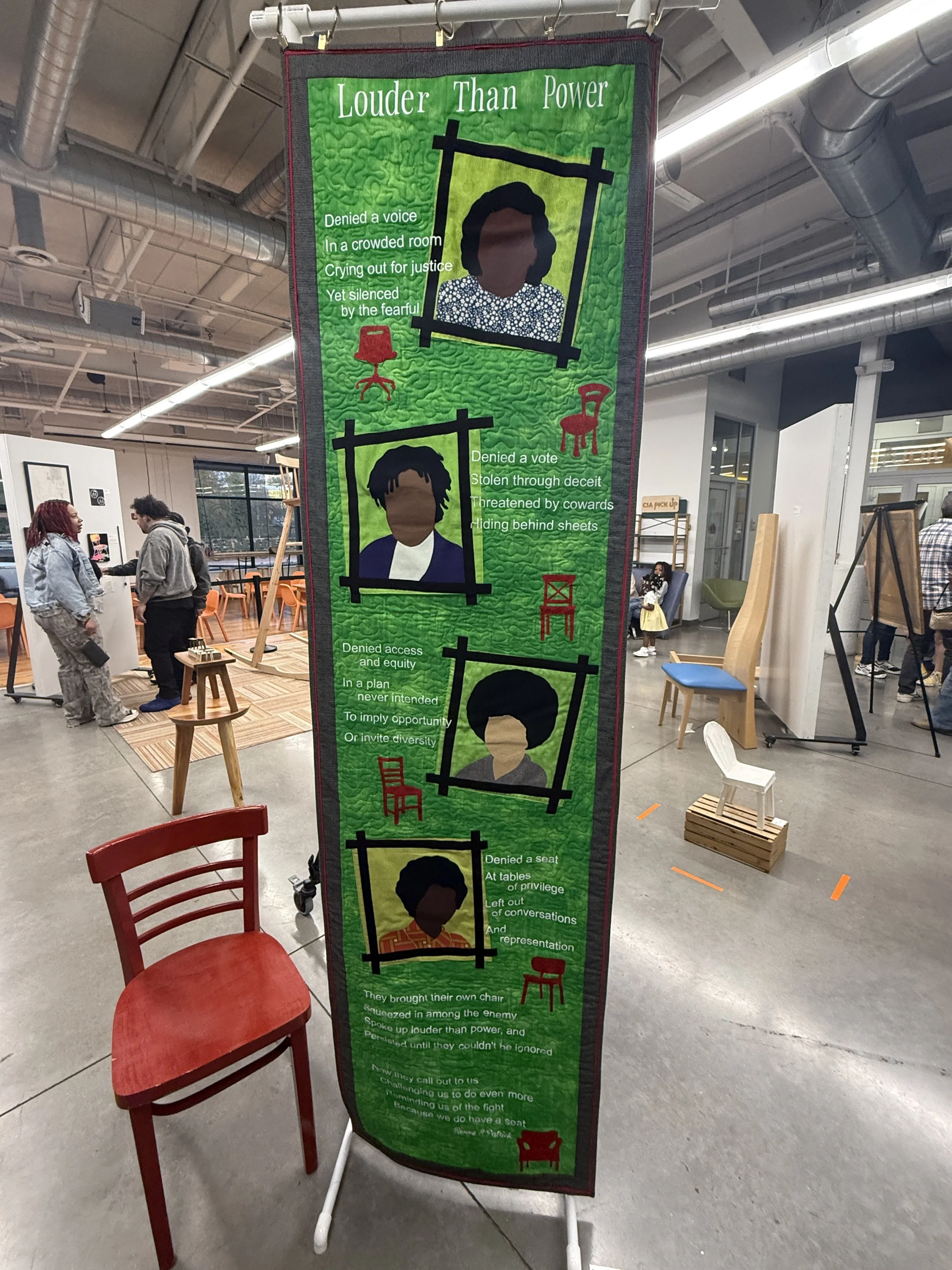 A tall green tapestry with illustrations of chairs and people, and text discussing themes of privilege, exclusion, and activism. The tapestry is displayed in an indoor space with chairs and people visible in the background.